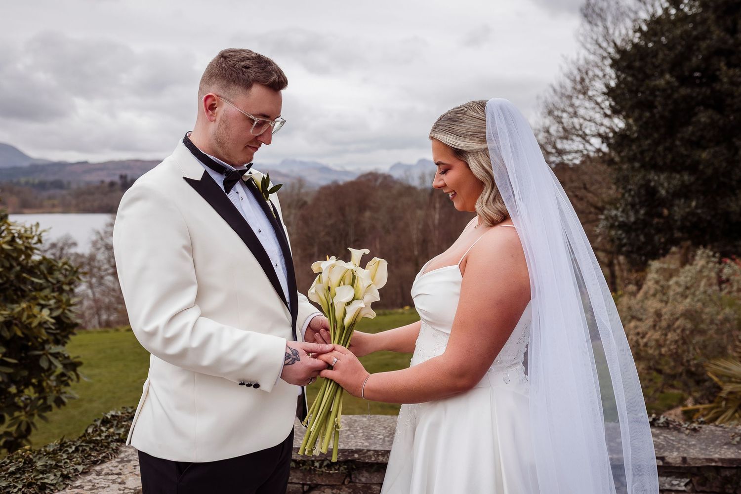 Groom in white tuxedo places ring on bride in wedding dress holding white calla lily bouquet outdoors.