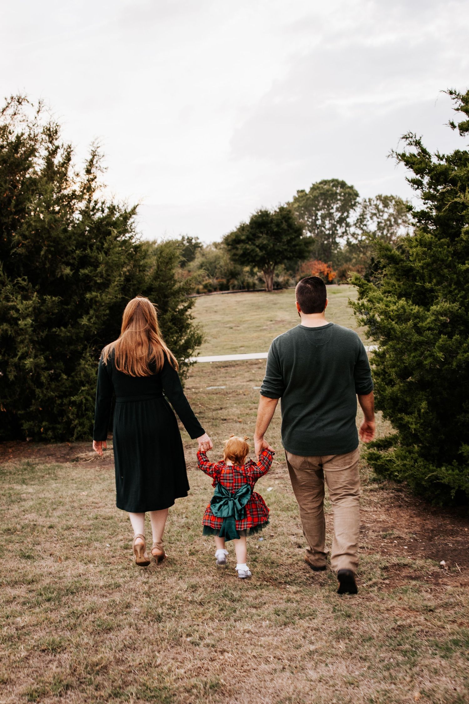 Family walking away holding hands with toddler in plaid dress between parents near evergreen trees.