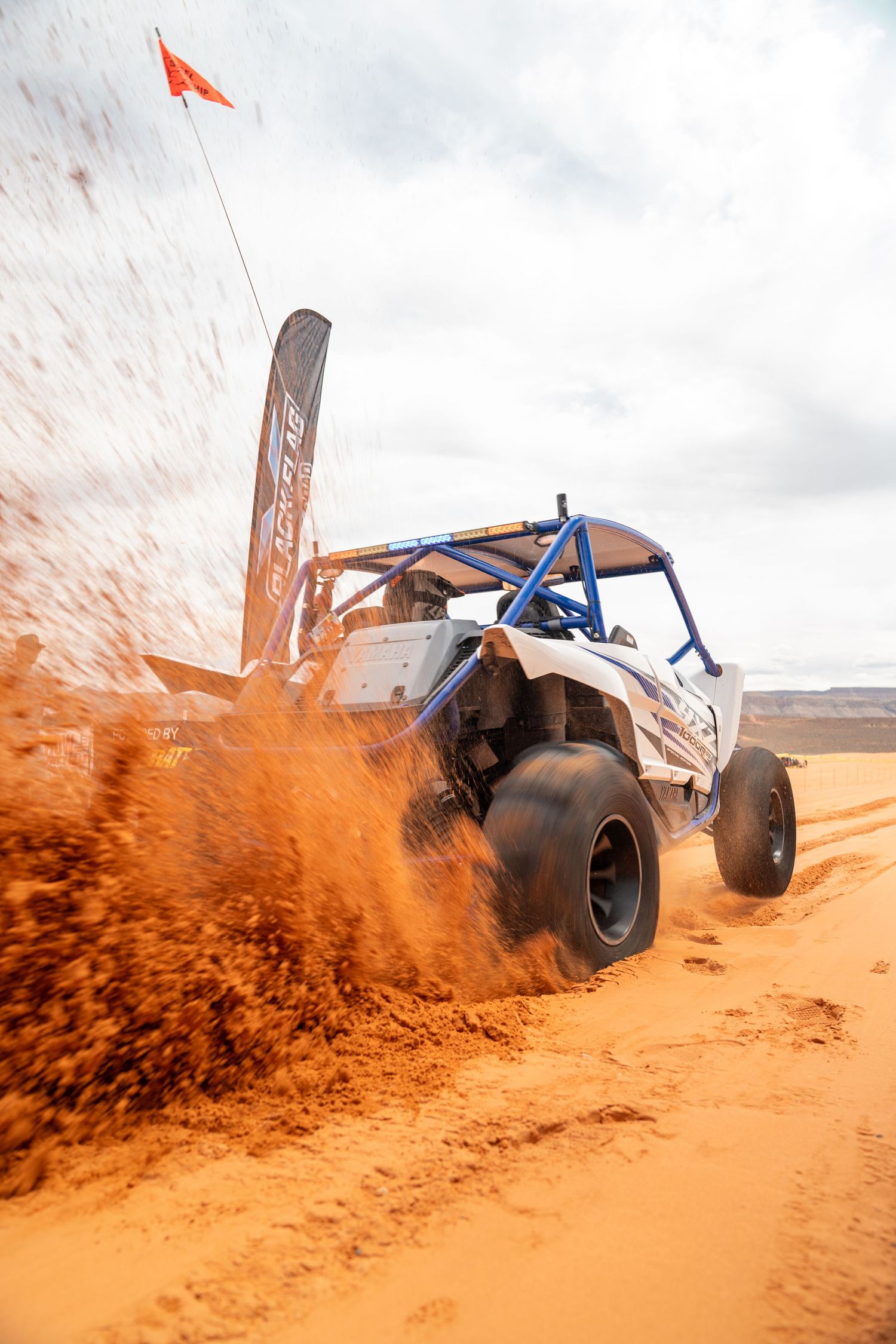 Off-road UTVs lined up at a desert racing event on red sand terrain under cloudy skies in a rugged landscape.