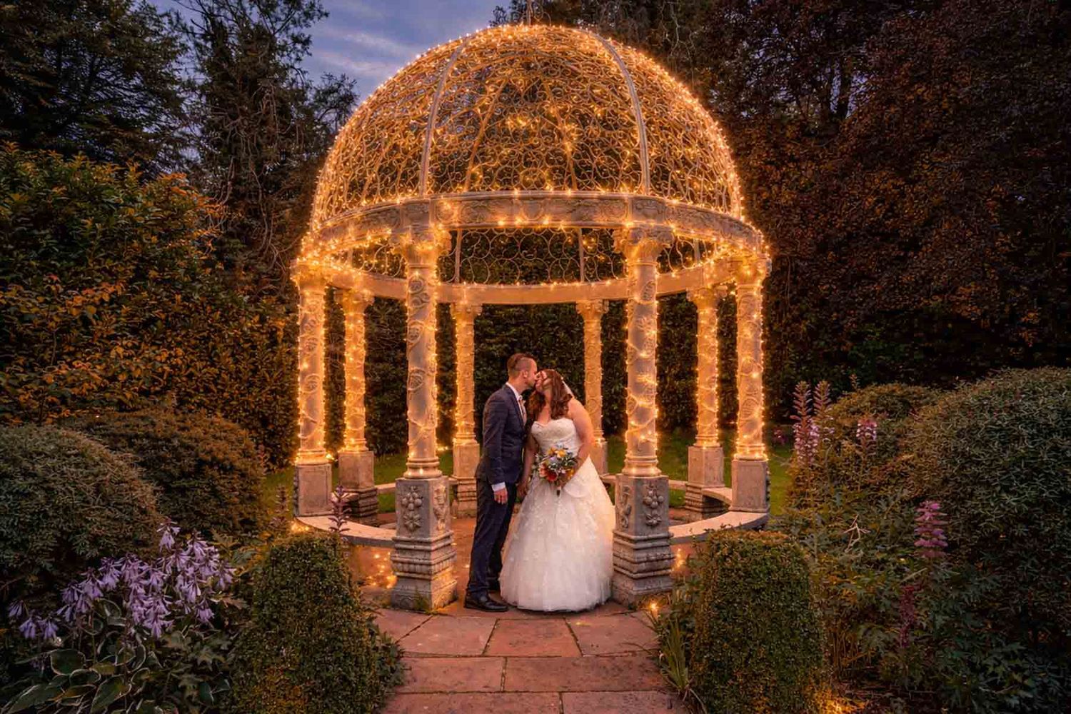 Bride and groom kissing under a fairy light gazebo in a romantic garden at dusk.