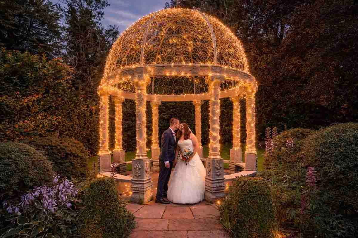 Bride and groom kissing under a fairy light gazebo in a romantic garden at dusk.