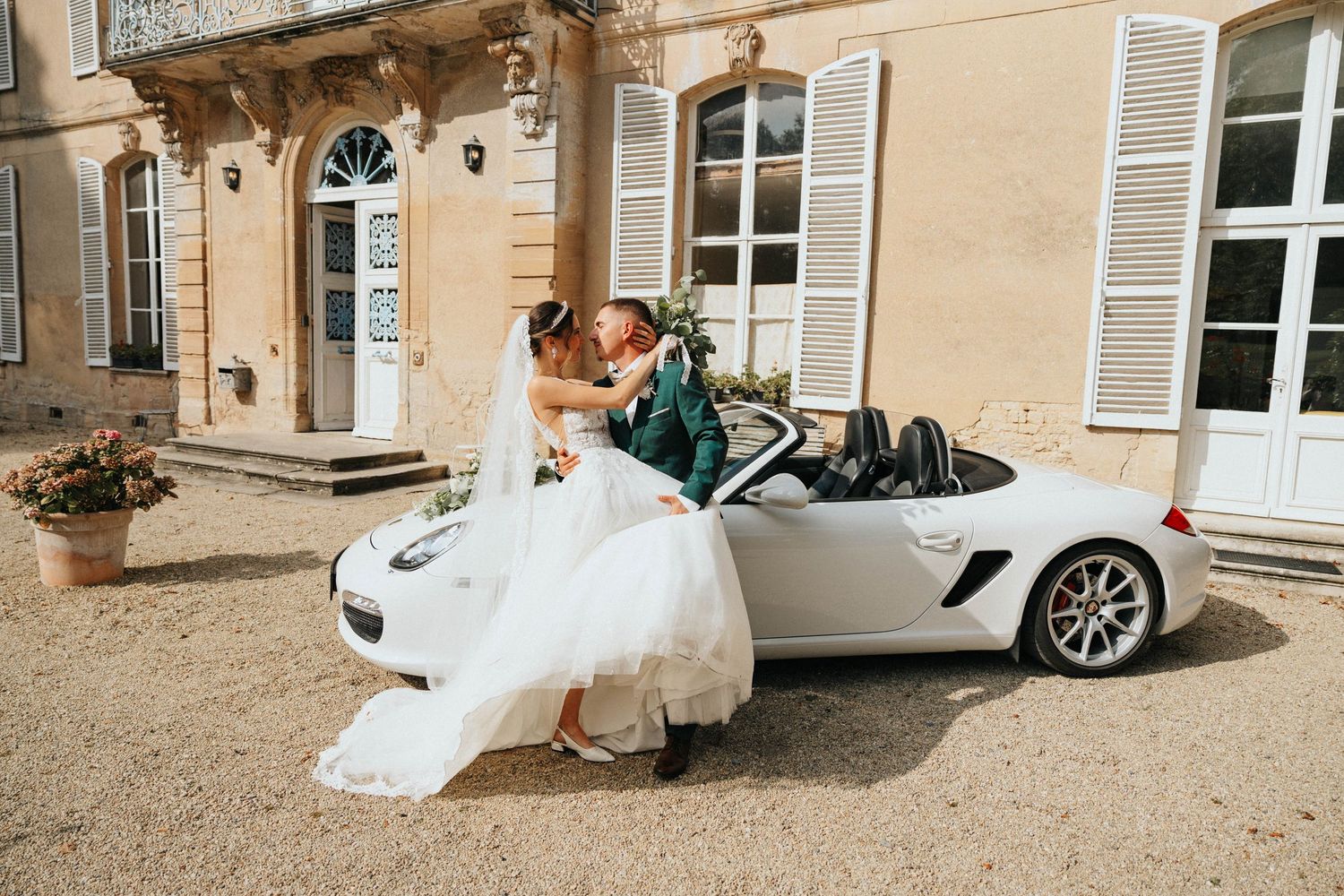 A bride and groom hold hands outside a historic stone church, she in a white gown holding a bouquet, he in a cream suit.