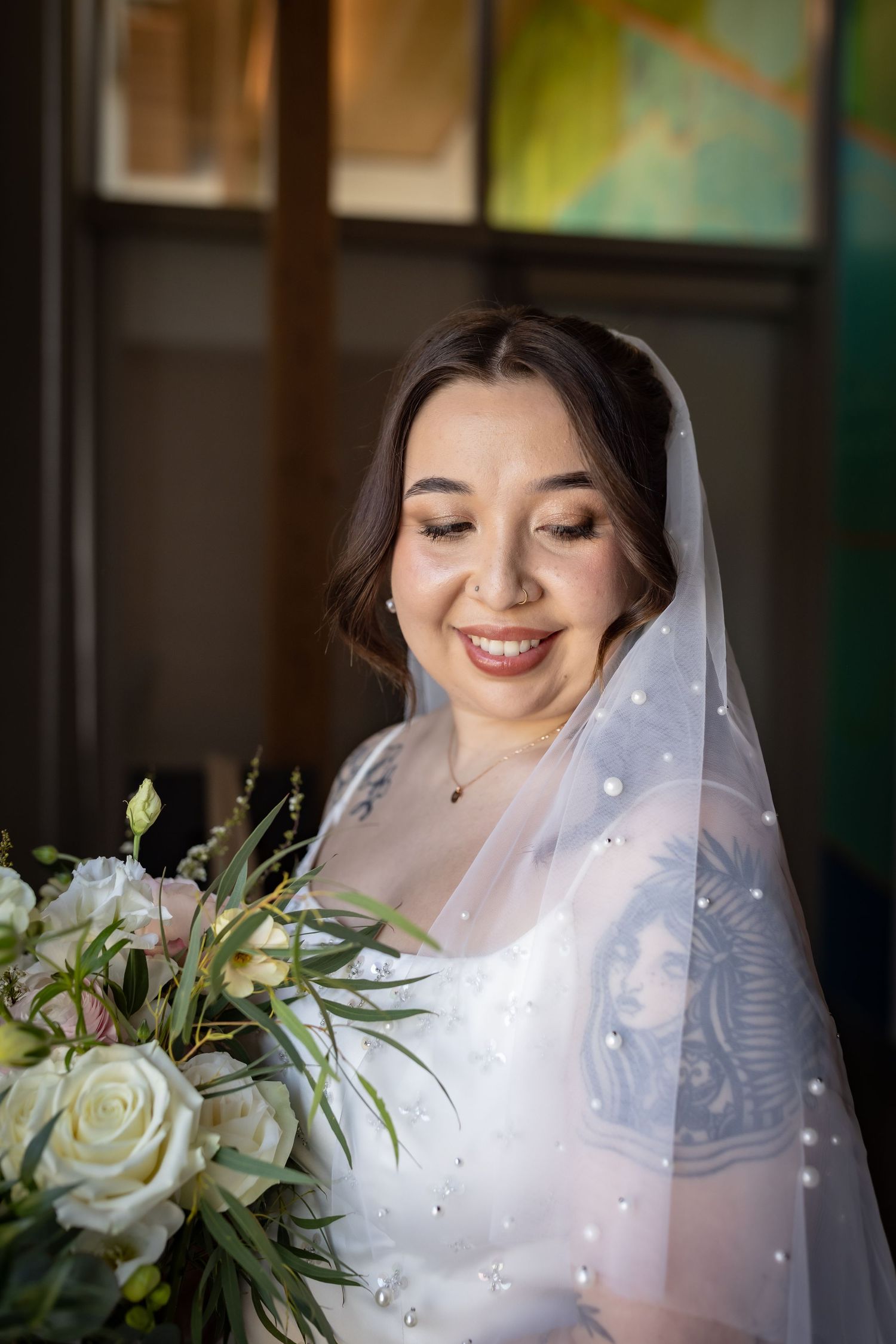 Smiling bride in white dress and pearl-adorned veil holds lush white rose bouquet in soft light.