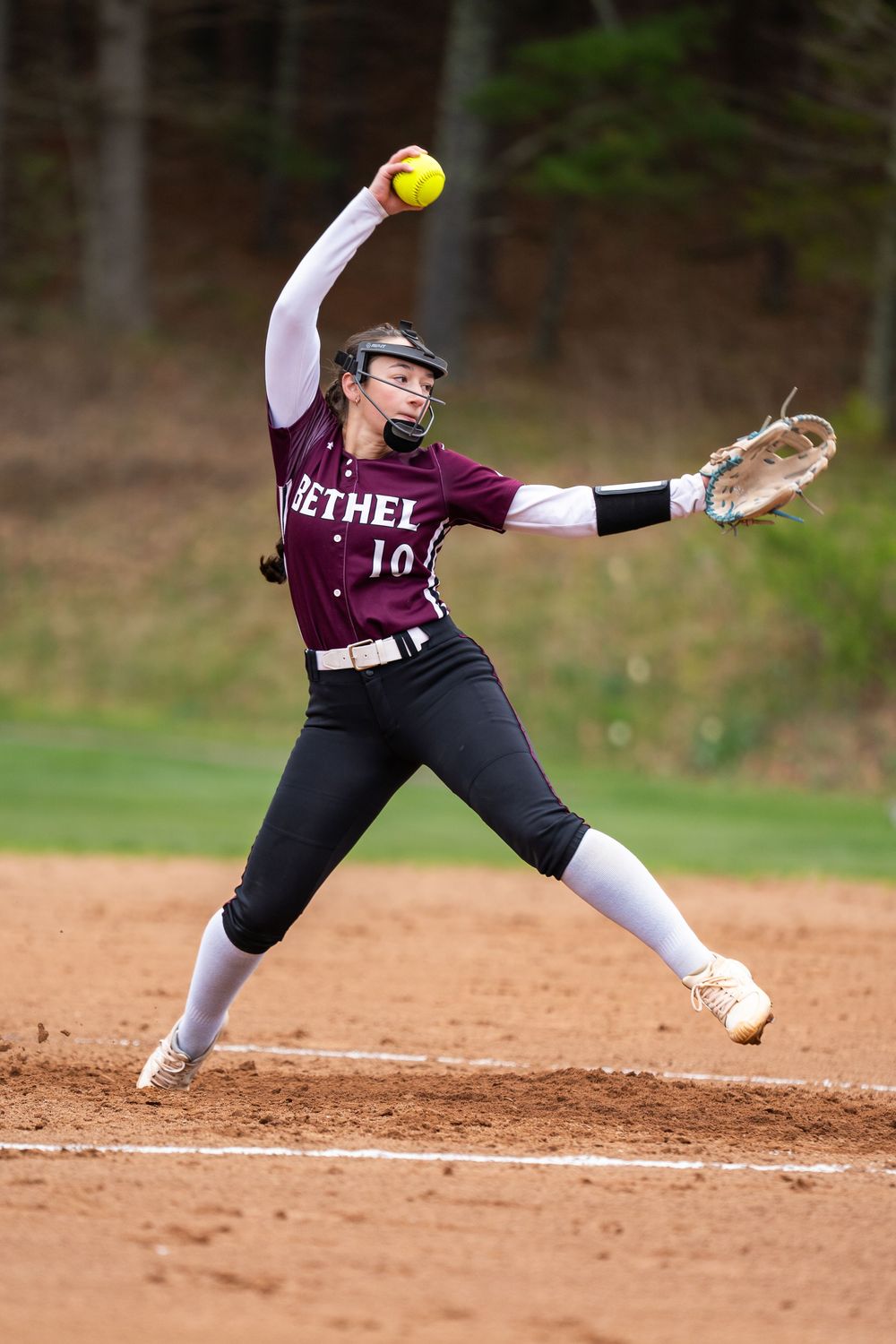 Bethel softball pitcher #10 winds up to deliver a pitch during a game on a dirt field.