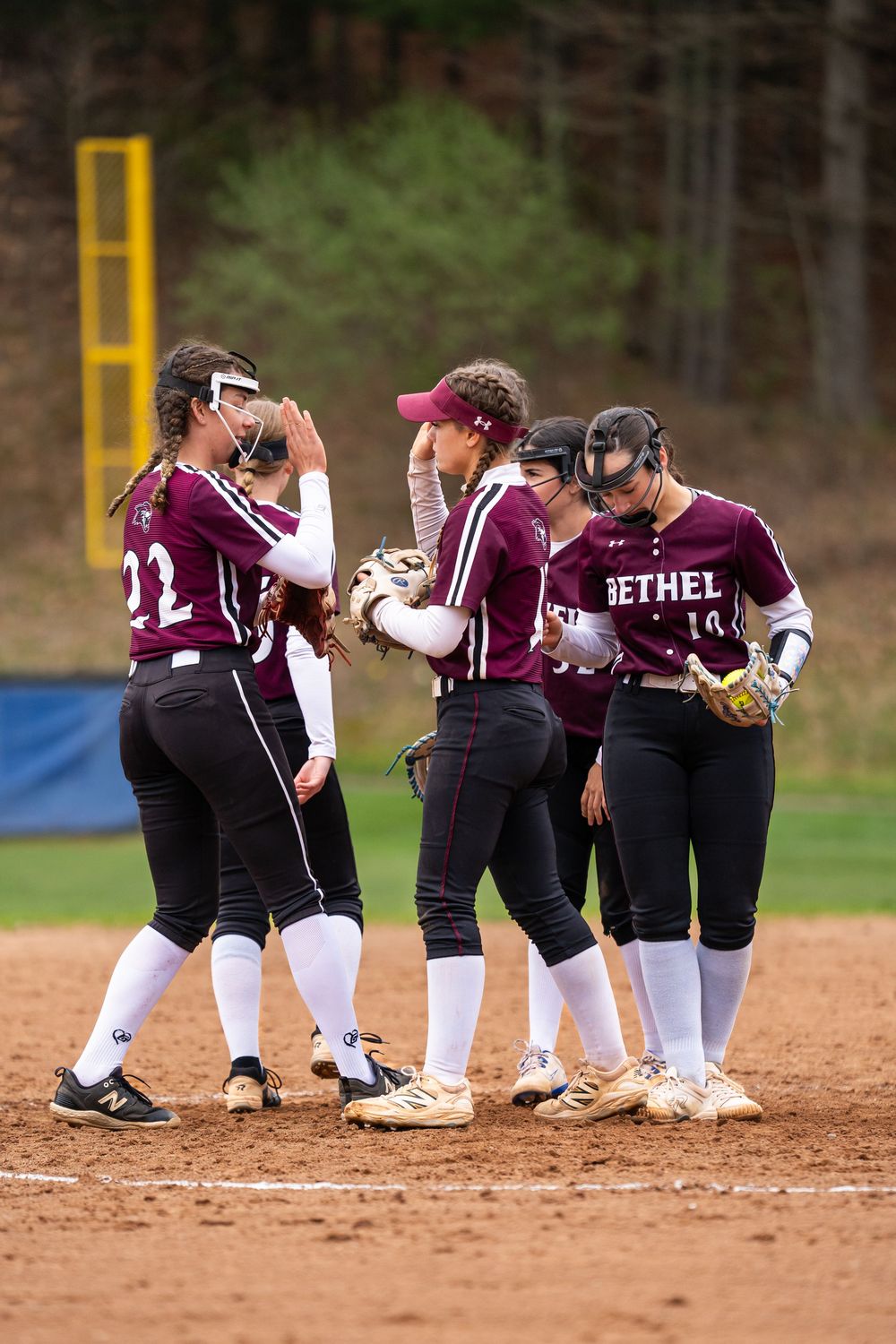 Bethel softball team players in maroon uniforms huddle together on the infield during a game.
