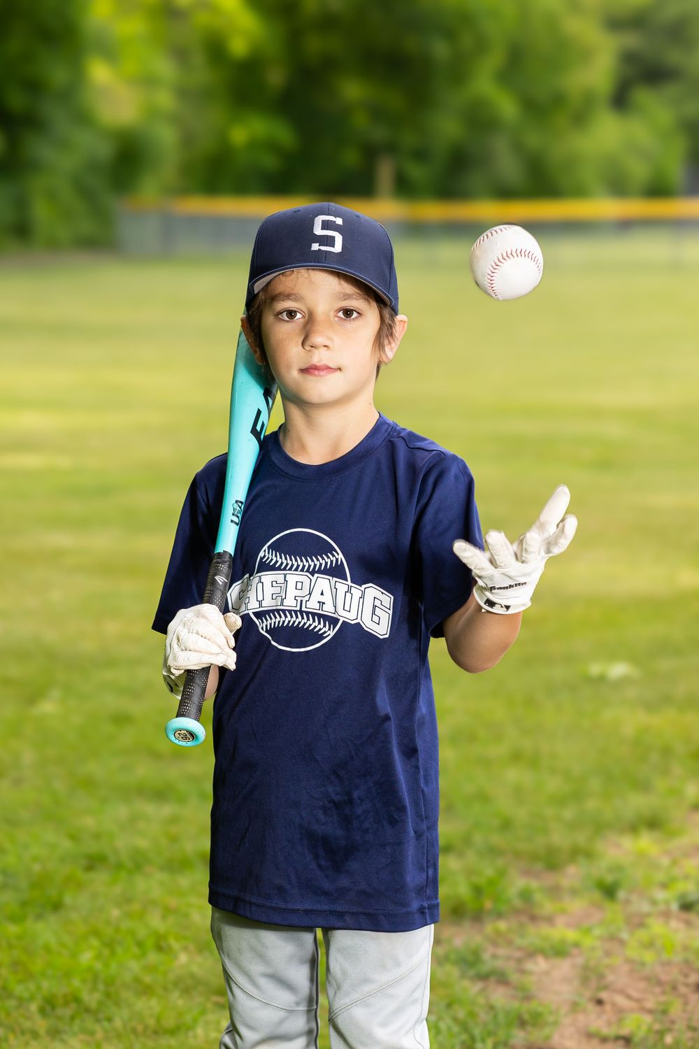 Young baseball player in navy uniform tossing a ball while holding a bat on a green field.