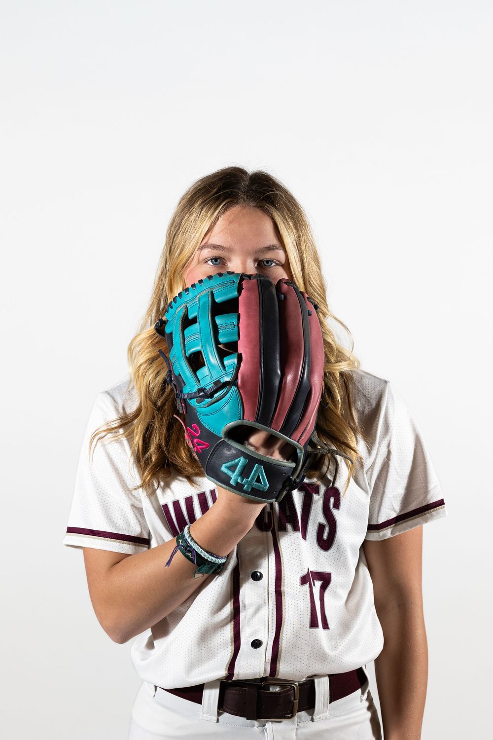 Female softball player in Wildcats uniform #17 holding a teal and pink fielding glove up to her face.