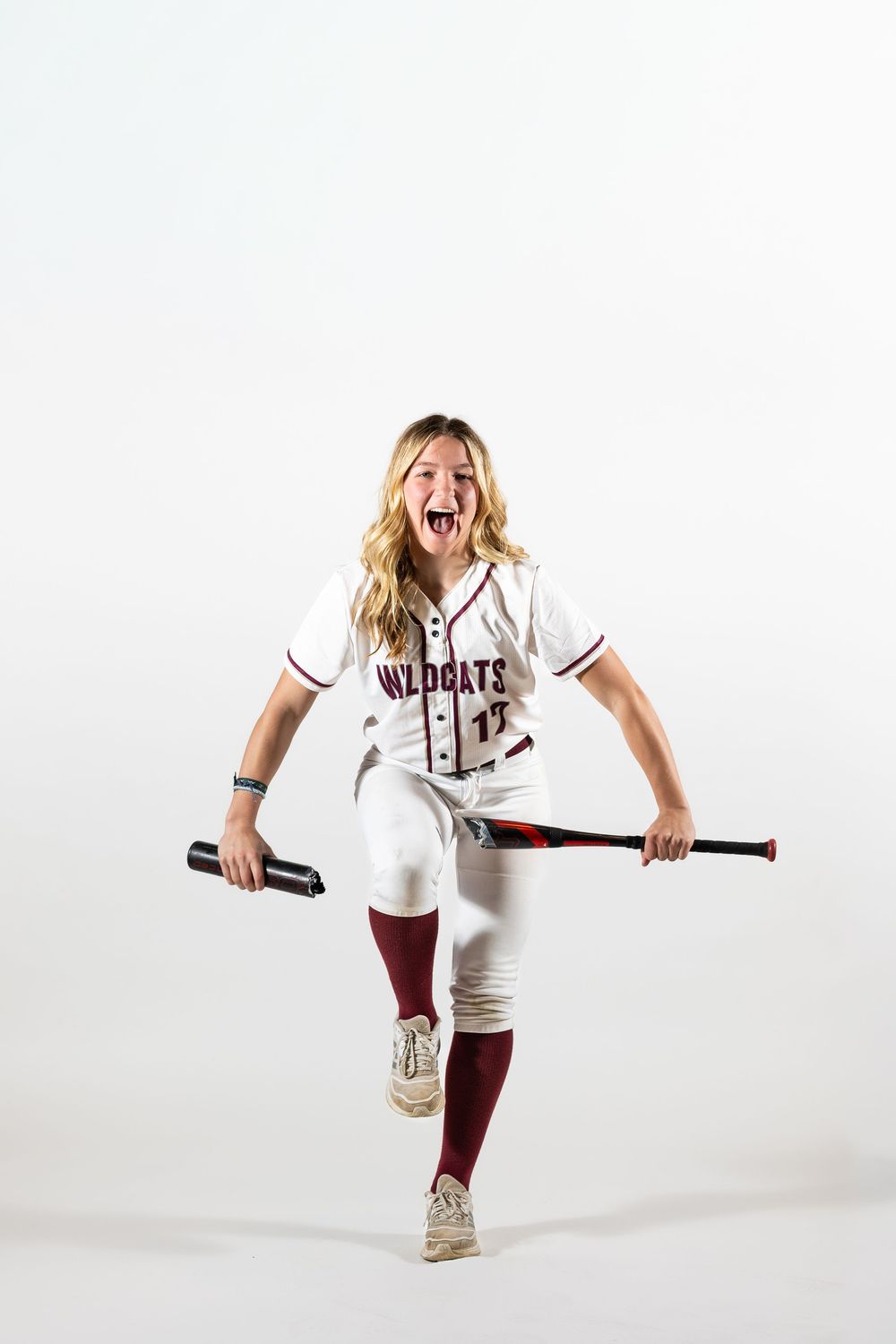 Energetic female Wildcats softball player #17 in white uniform holds bat, wearing maroon socks, posing on white background.