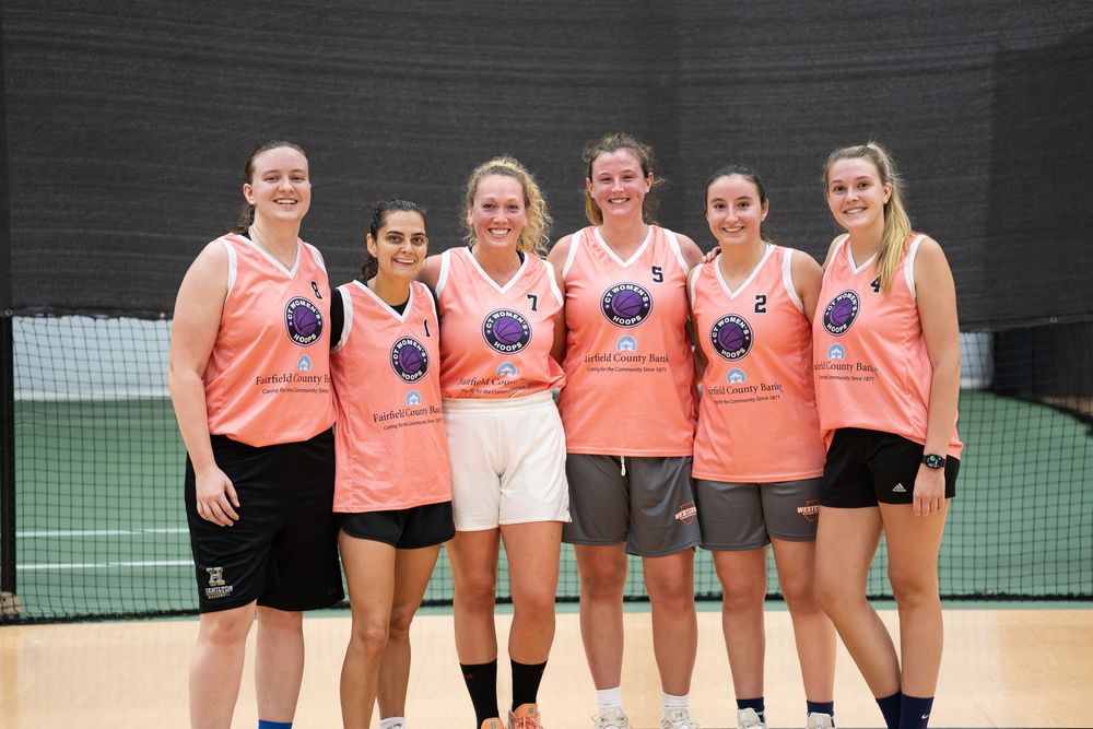Six women in matching pink jerseys pose together smiling on an indoor tennis court after a team sports event.
