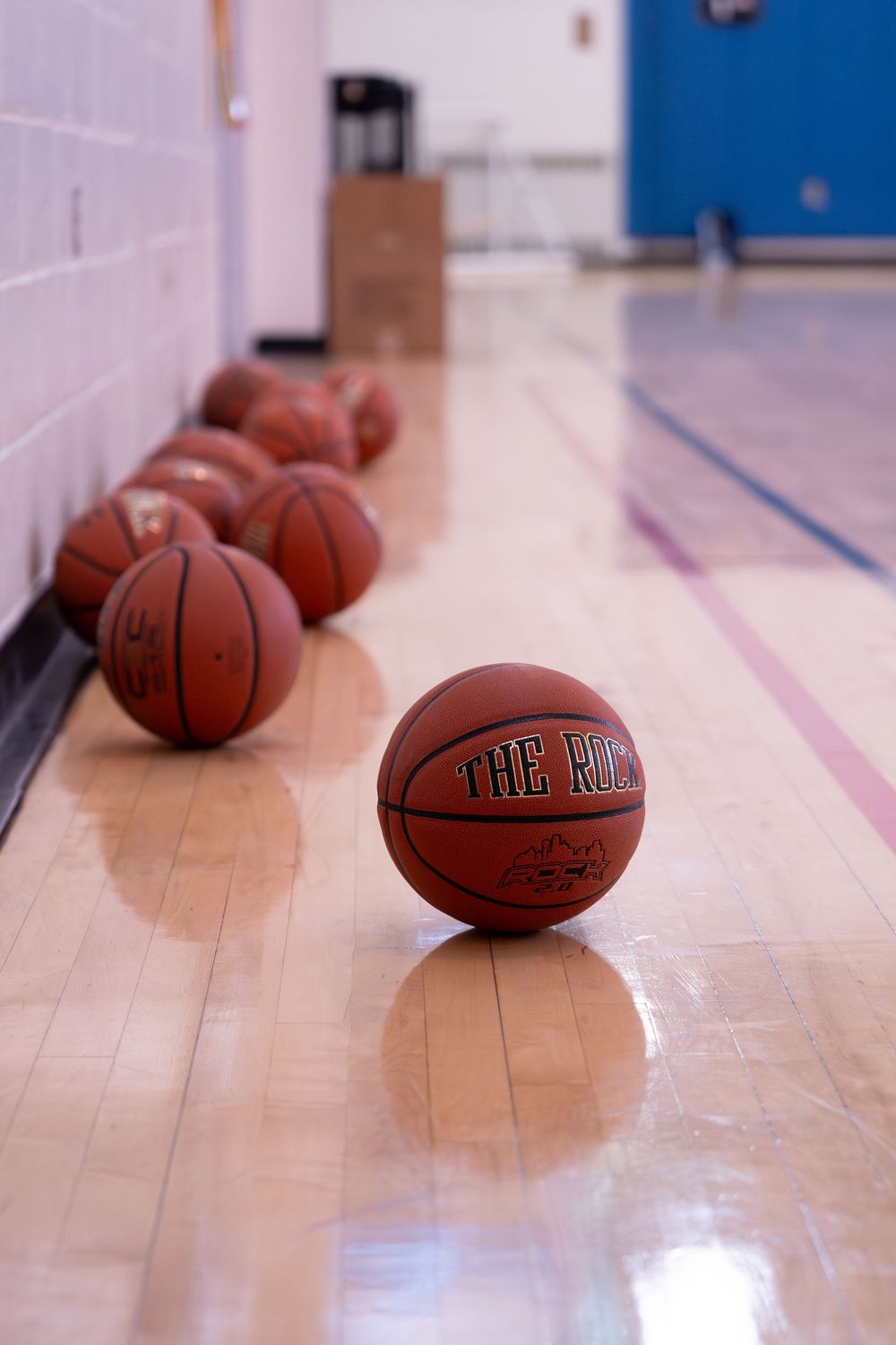 Multiple basketballs labeled The Rock lined up on a shiny hardwood gymnasium floor along a white brick wall.