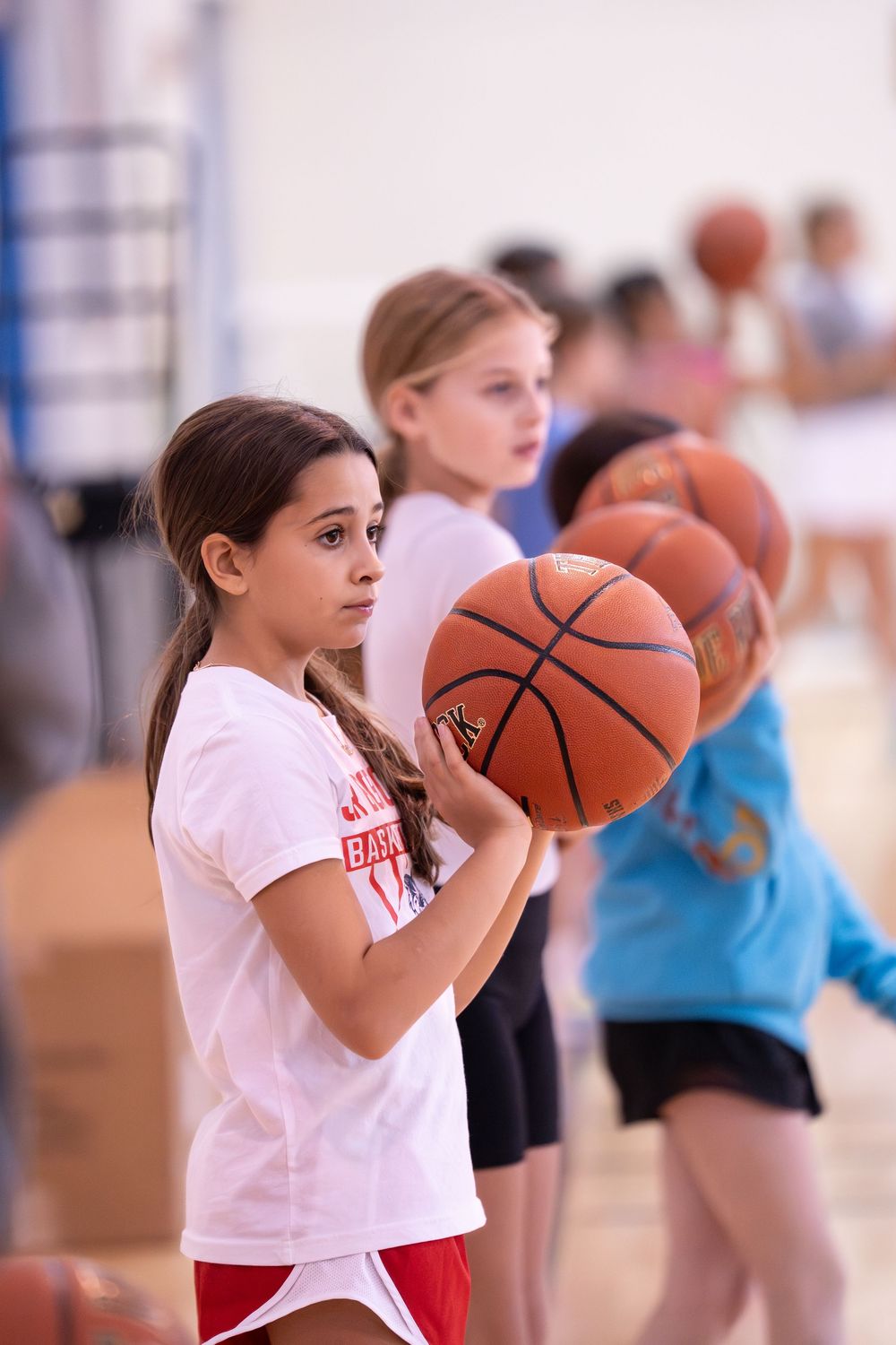 Girls practicing basketball drills in a gymnasium, holding basketballs in a line during a youth sports training session.