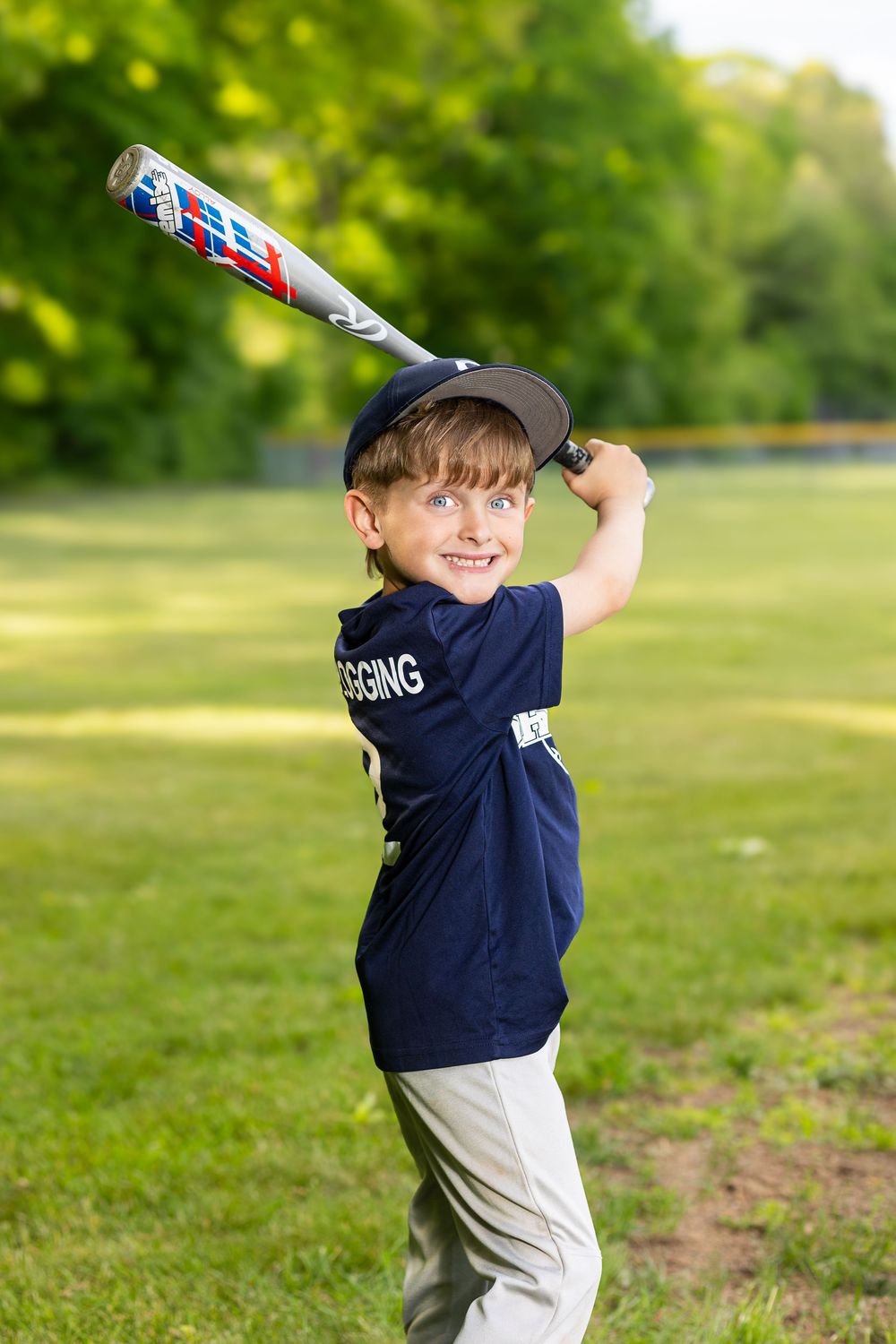 Young boy in navy baseball uniform swinging a bat on a green field, smiling during youth baseball practice.