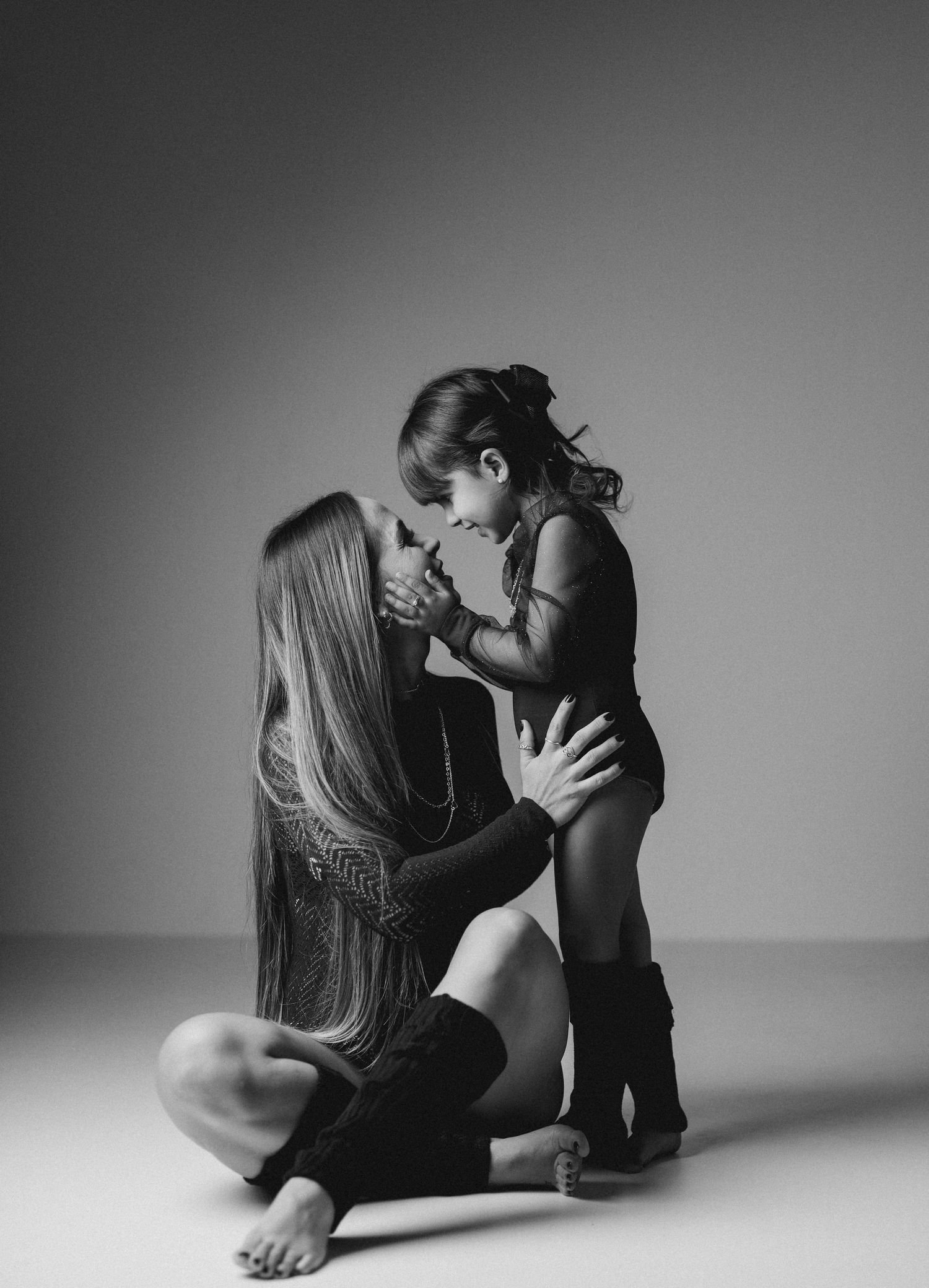 Black and white photo of a mother sitting cross-legged on the floor tenderly holding her young child's hands.