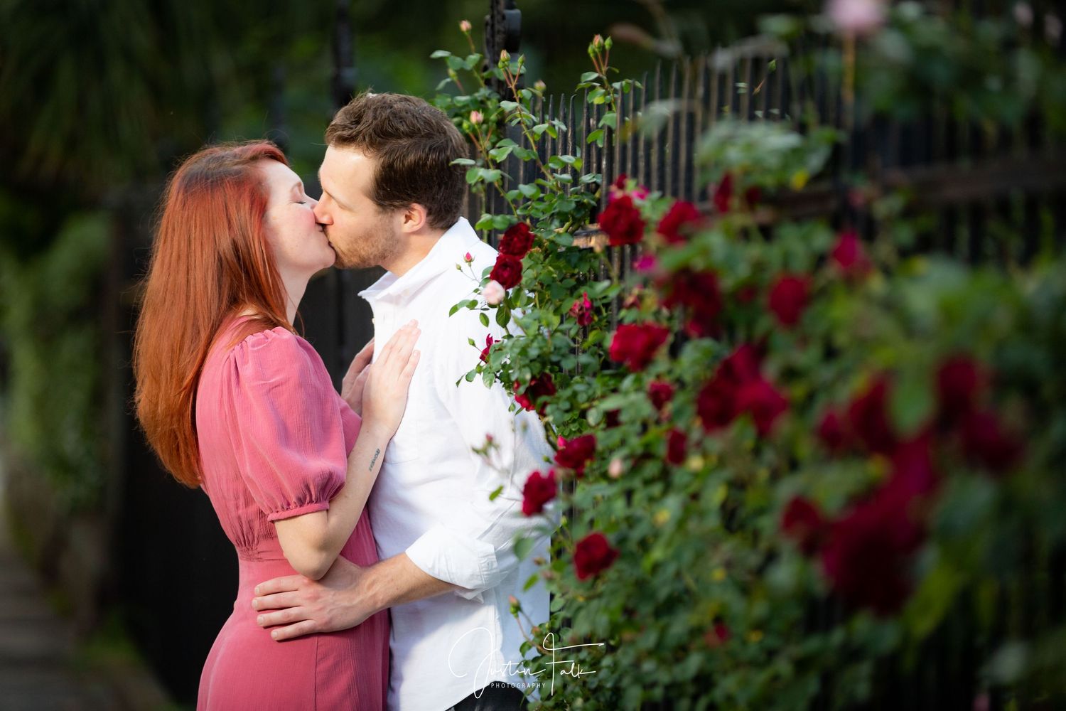 Couple sharing a romantic kiss beside blooming red roses in a lush garden setting.