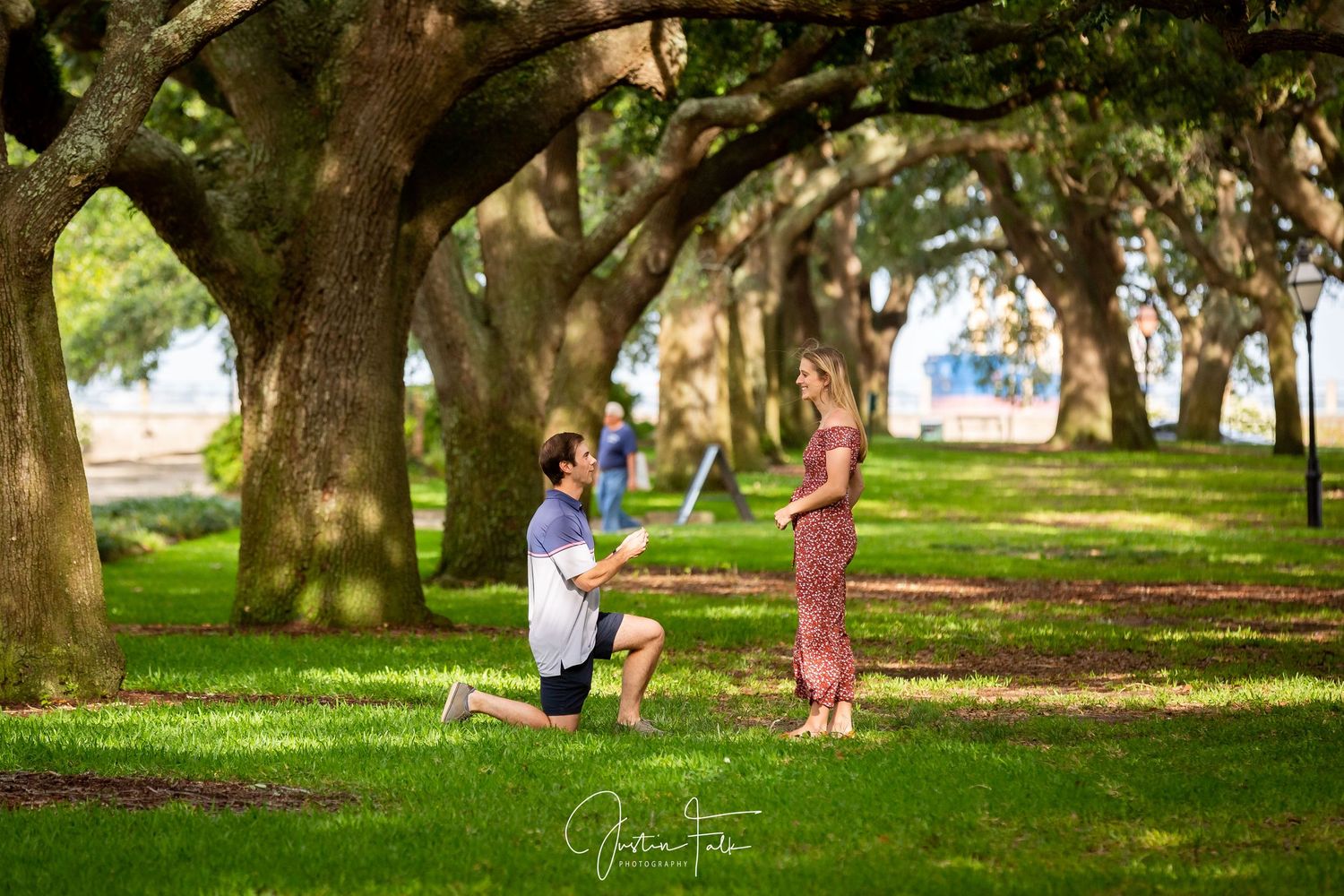 Man proposing on one knee to his partner in a scenic tree-lined park.