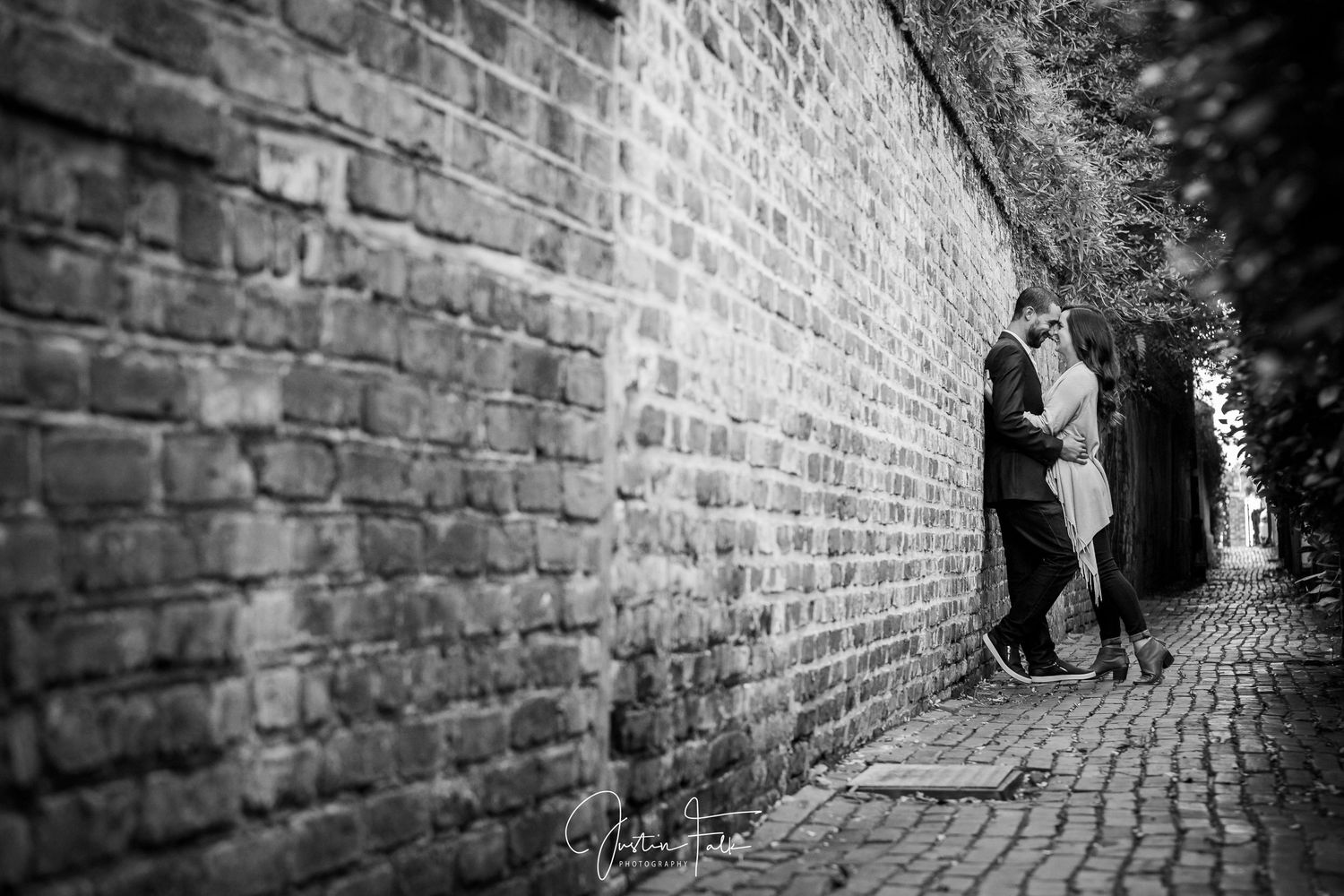Black and white photo of a couple sharing a romantic kiss against a rustic brick wall on a cobblestone alley.