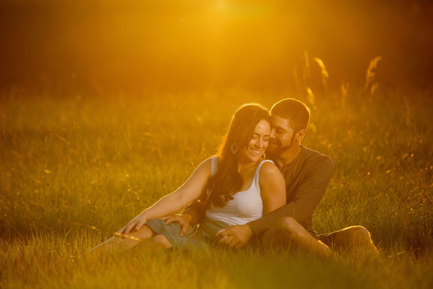 Couple sitting together in golden sunset light on a lush green field, sharing a romantic intimate moment.