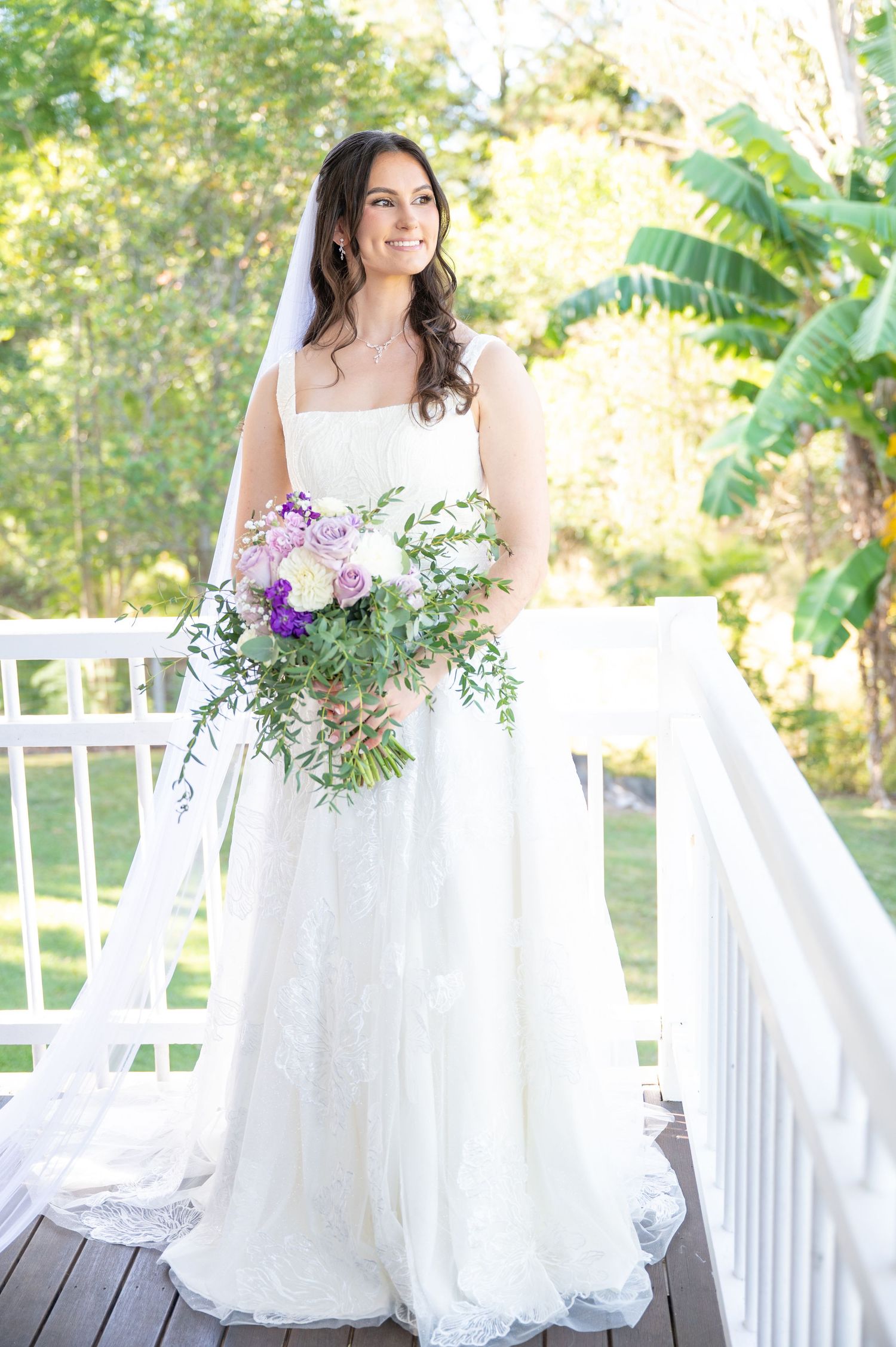 Bride in white lace gown holding purple and white bouquet on deck.
