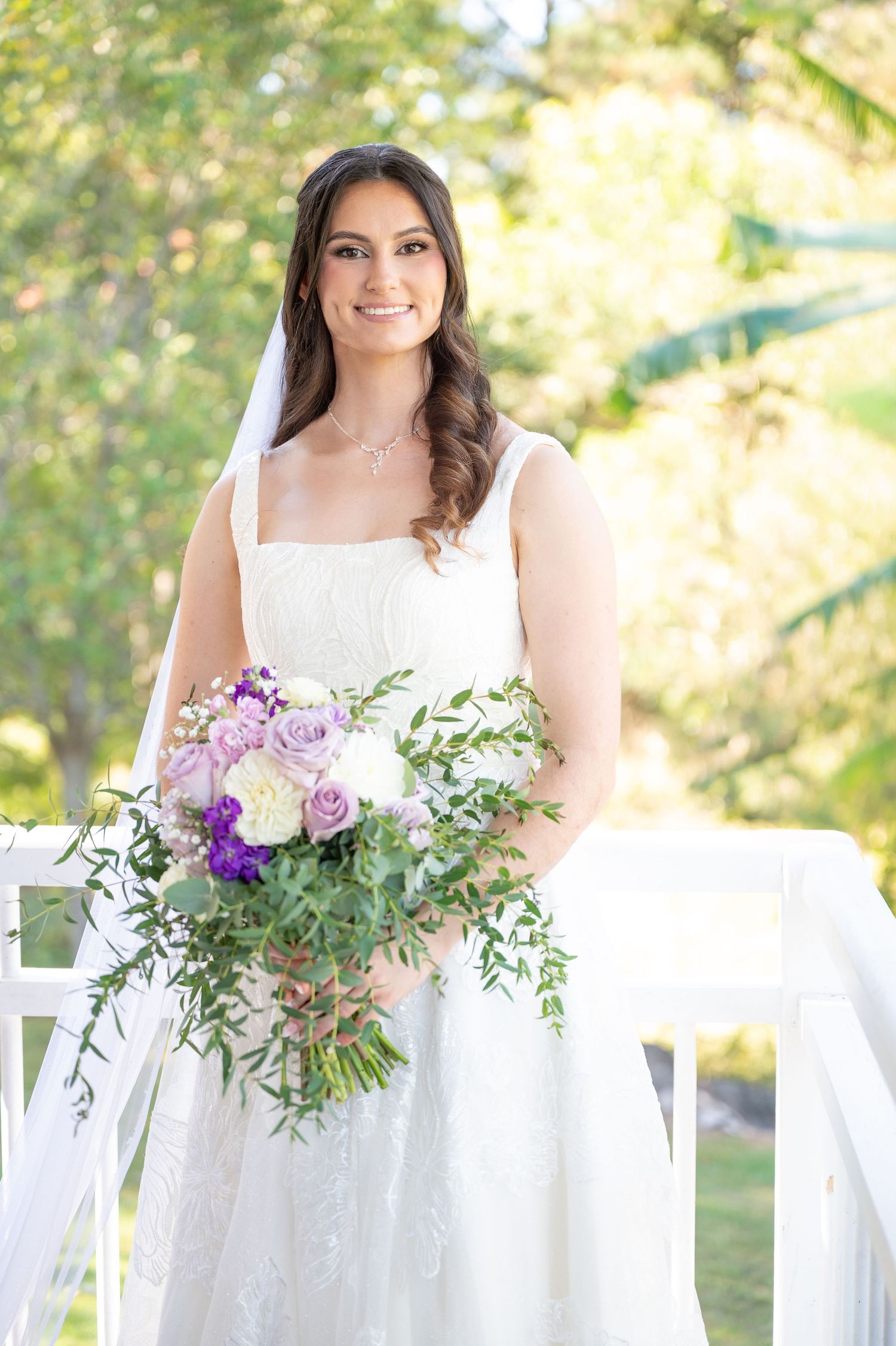 Smiling bride in white lace dress holding purple bouquet on white balcony.