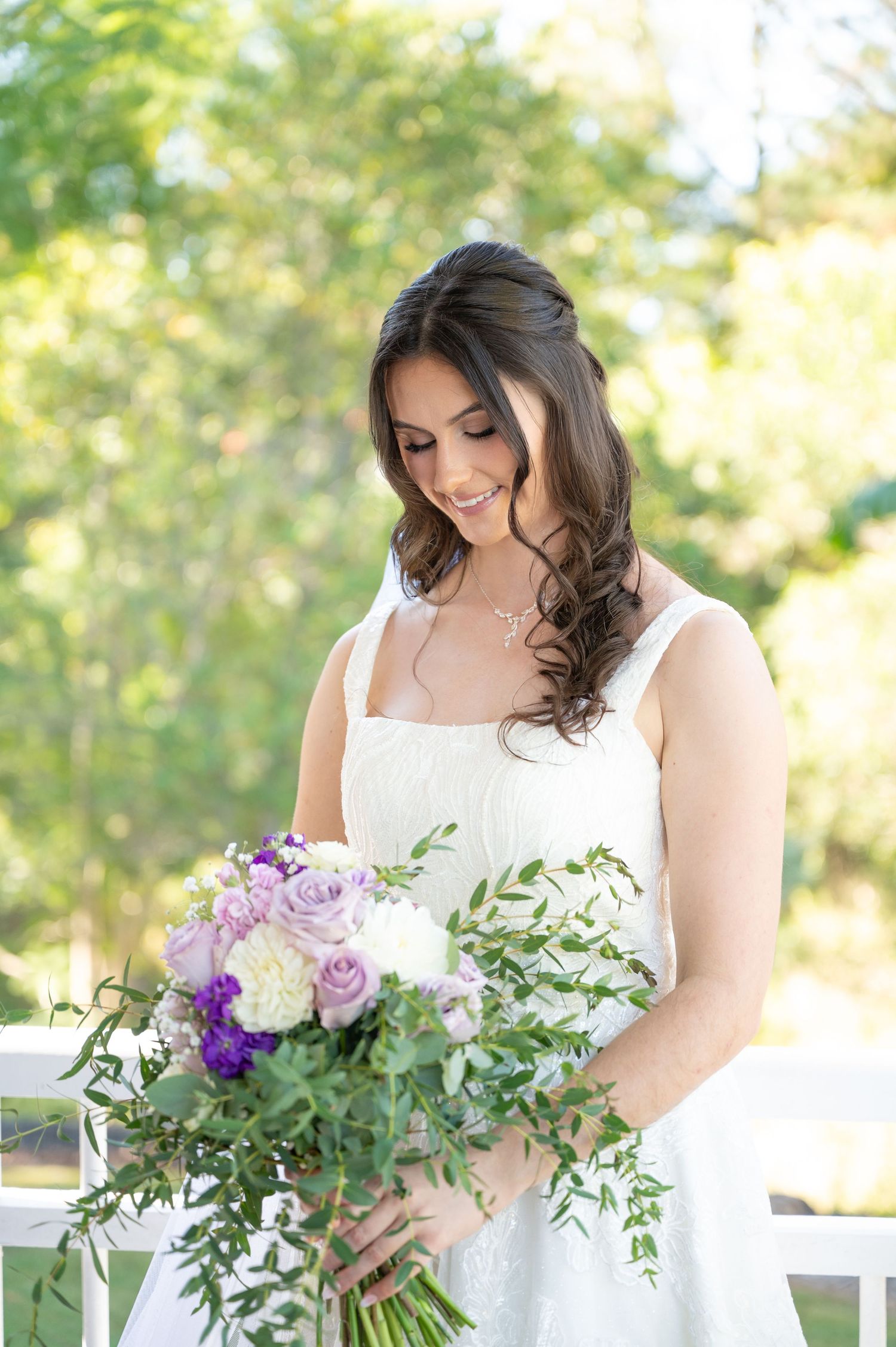 Bride in white lace gown admiring purple and white bouquet outdoors.