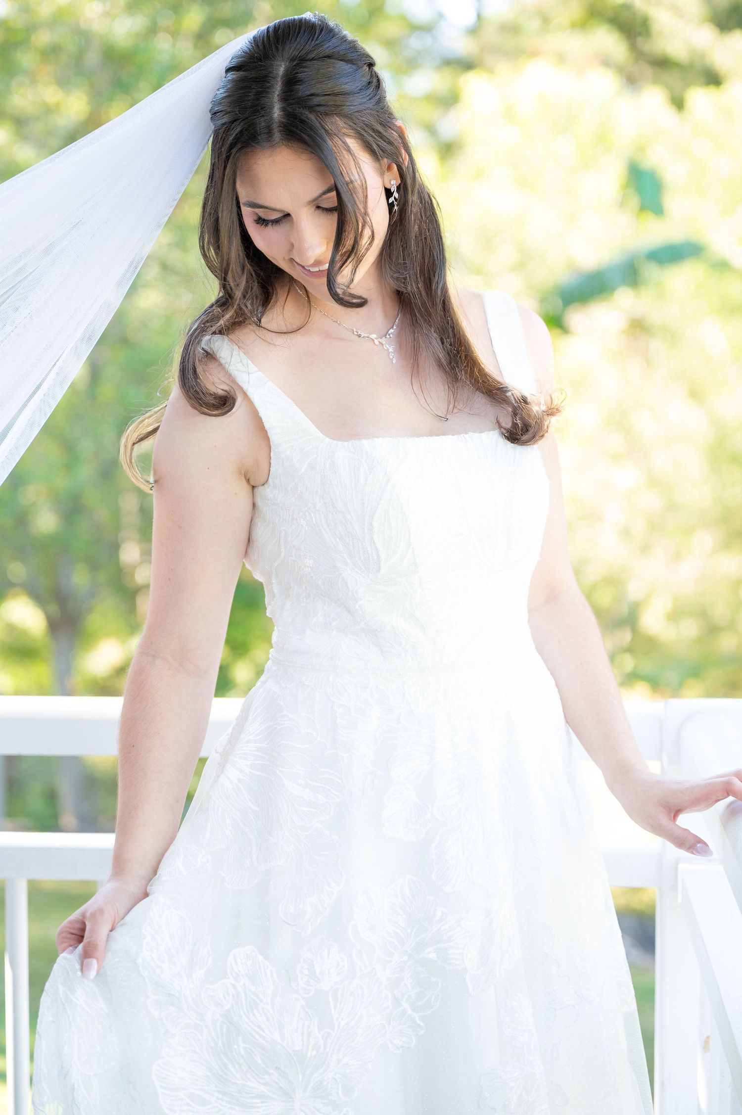 Bride in elegant white lace gown with veil on sunlit white balcony.