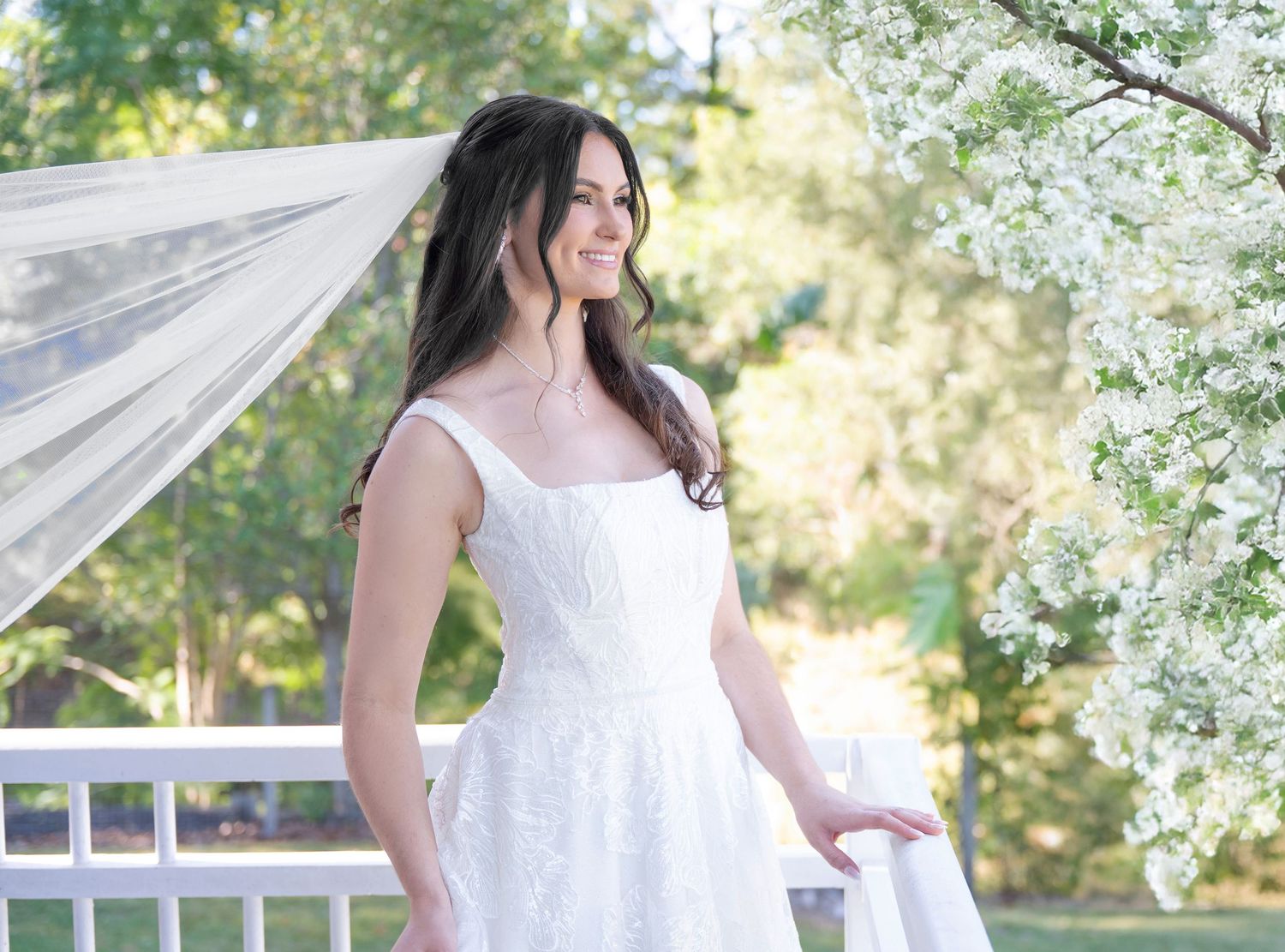 Elegant bride in white wedding dress standing near blooming white flowers in a sunny garden.