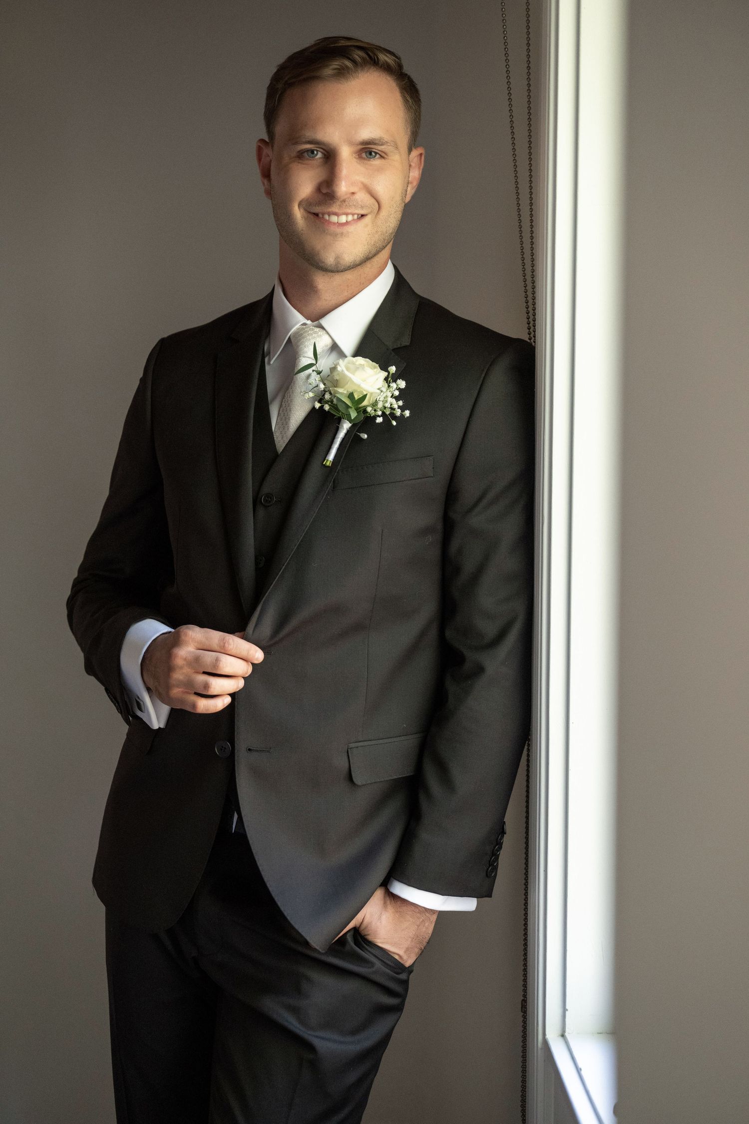 Smiling groom in black suit with white boutonniere standing by window.