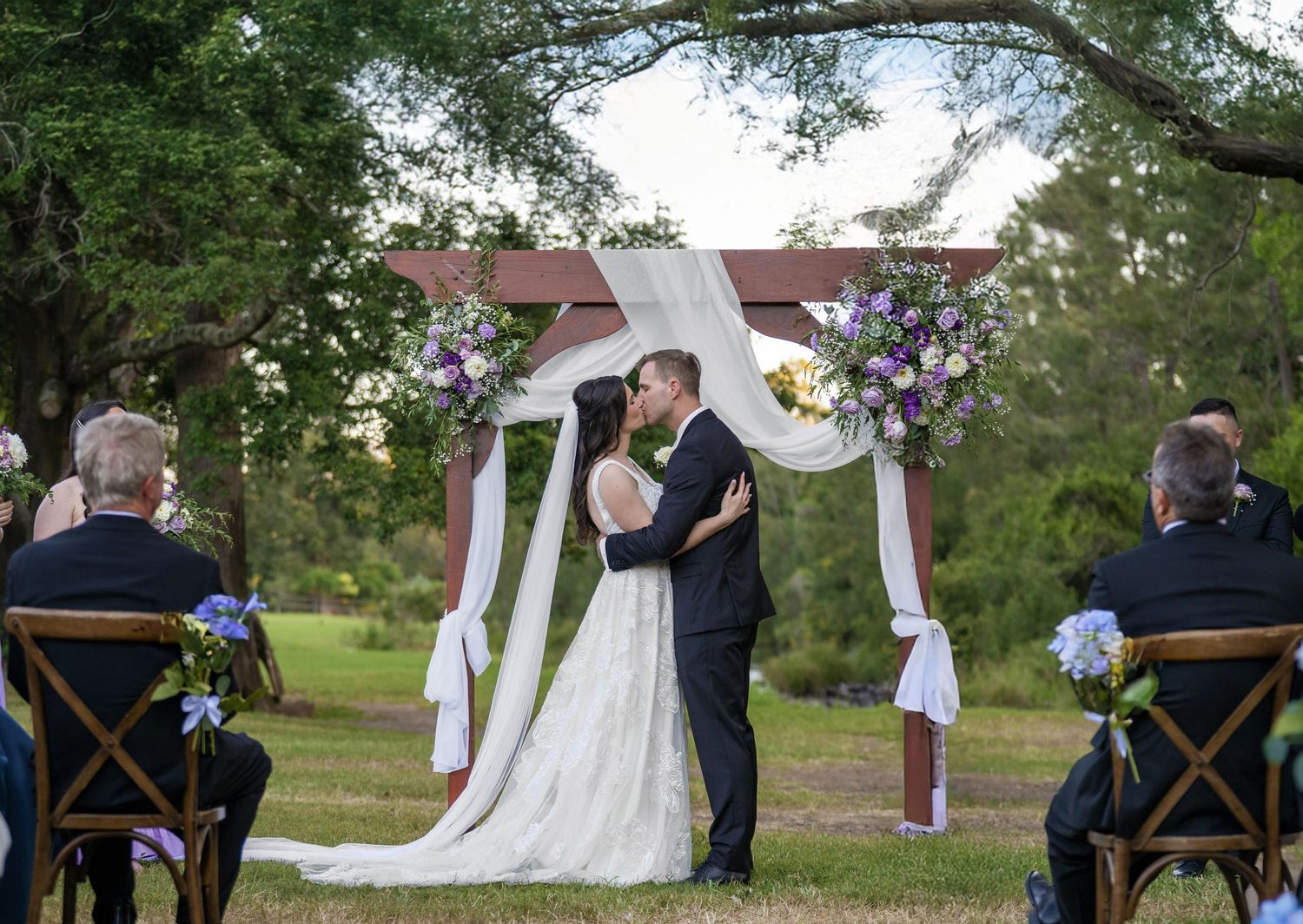 Bride and groom sharing a kiss at a floral decorated outdoor wedding ceremony arch at sunset.