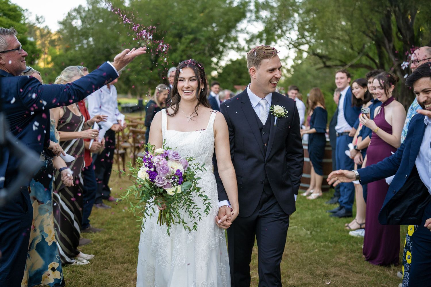 Happy bride and groom walking through confetti shower surrounded by cheering wedding guests outdoors.