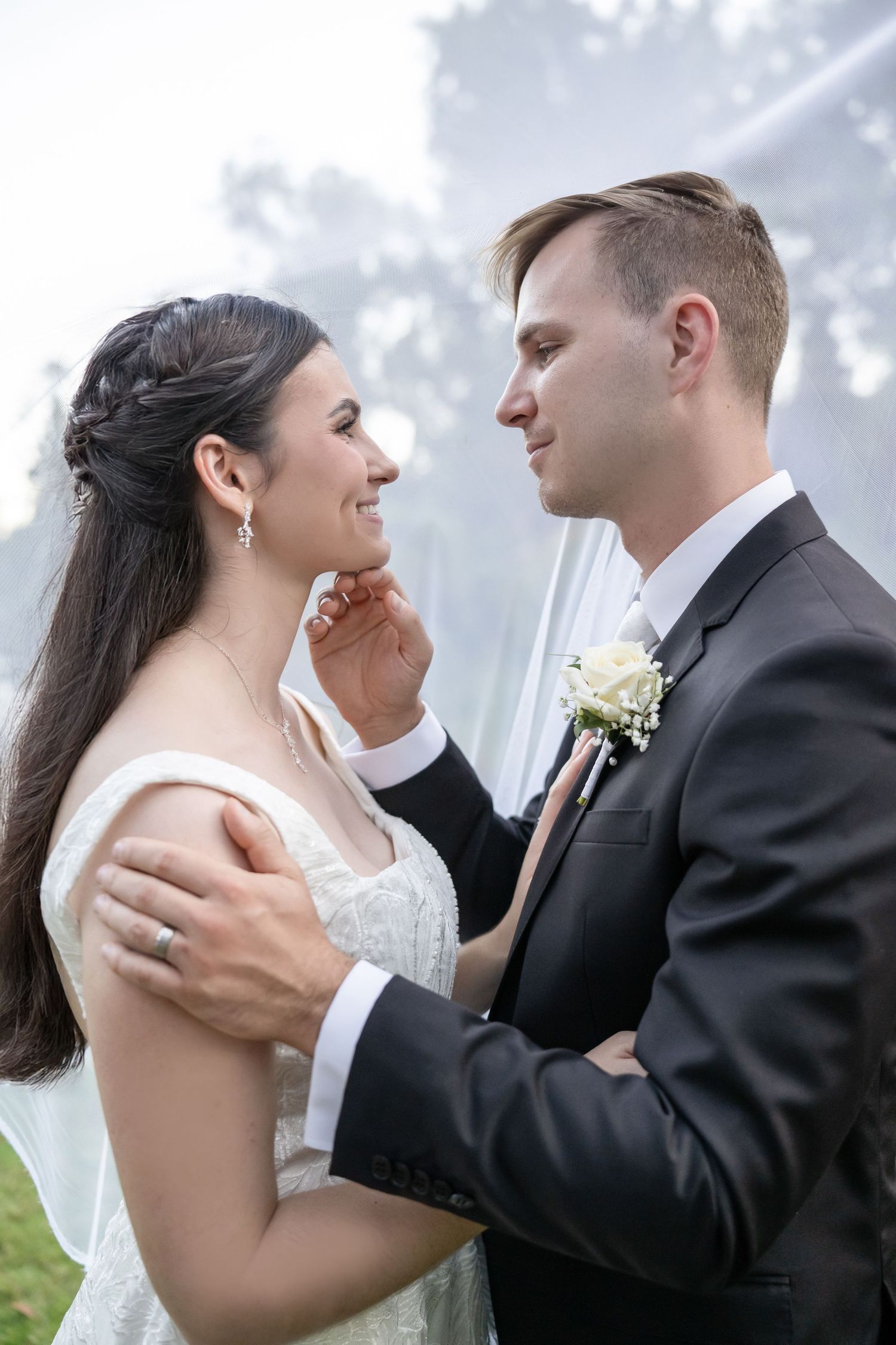 Groom tenderly touching bride's face as they gaze lovingly at each other outdoors.