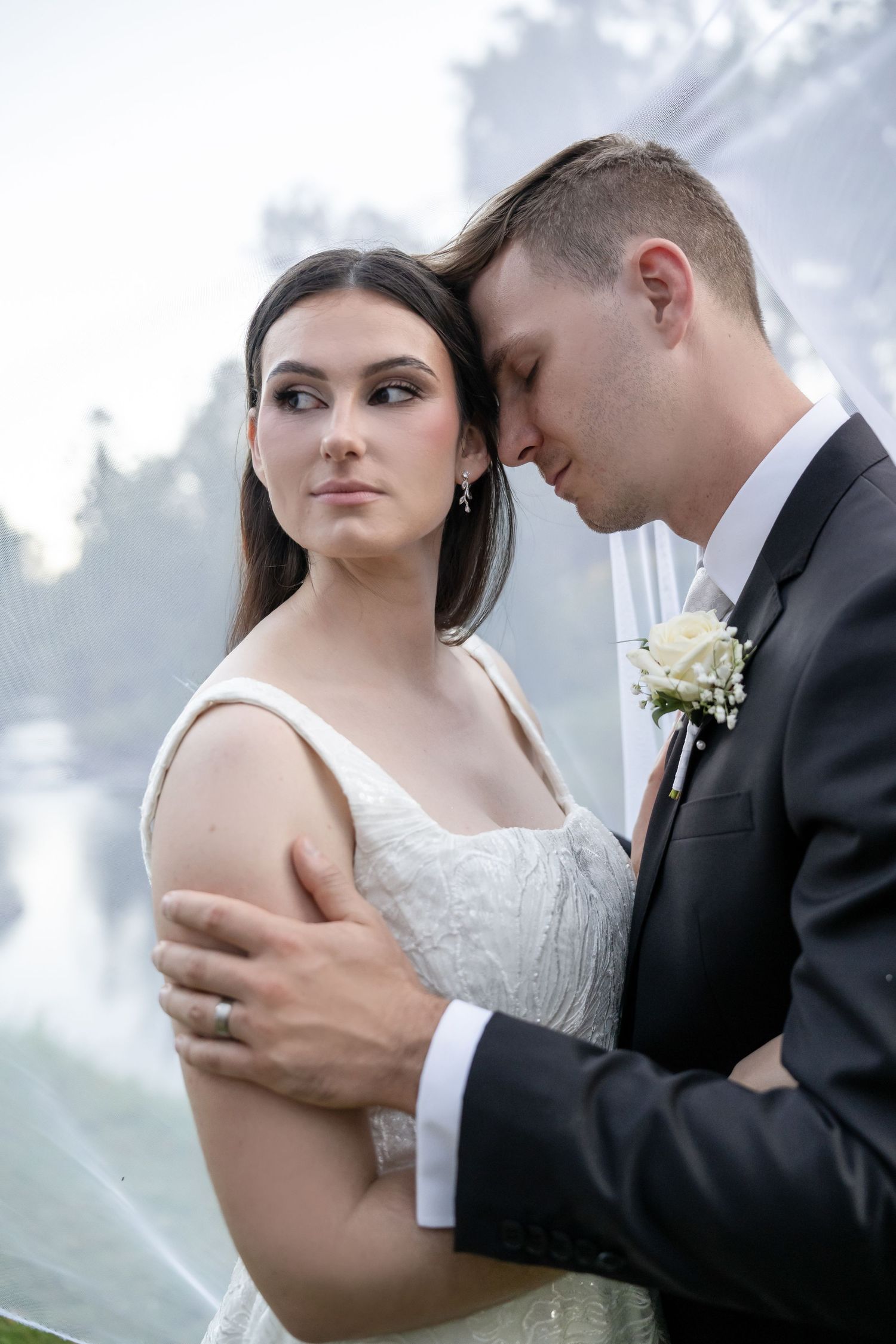 Elegant bride in white lace gown leaning against groom in black suit with white boutonniere.