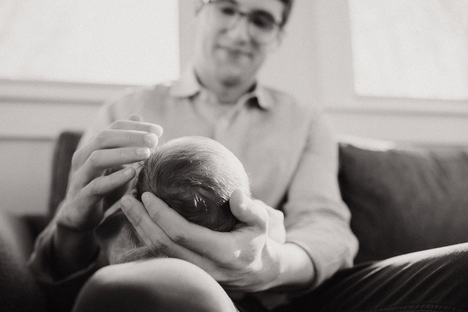 Black and white photo of a smiling man holding a newborn baby in his hands while seated on a couch.