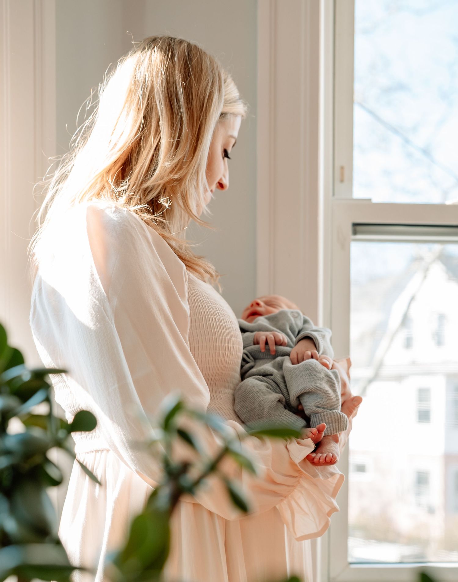 A blonde mother in a white dress tenderly holds her newborn baby by a sunlit window.