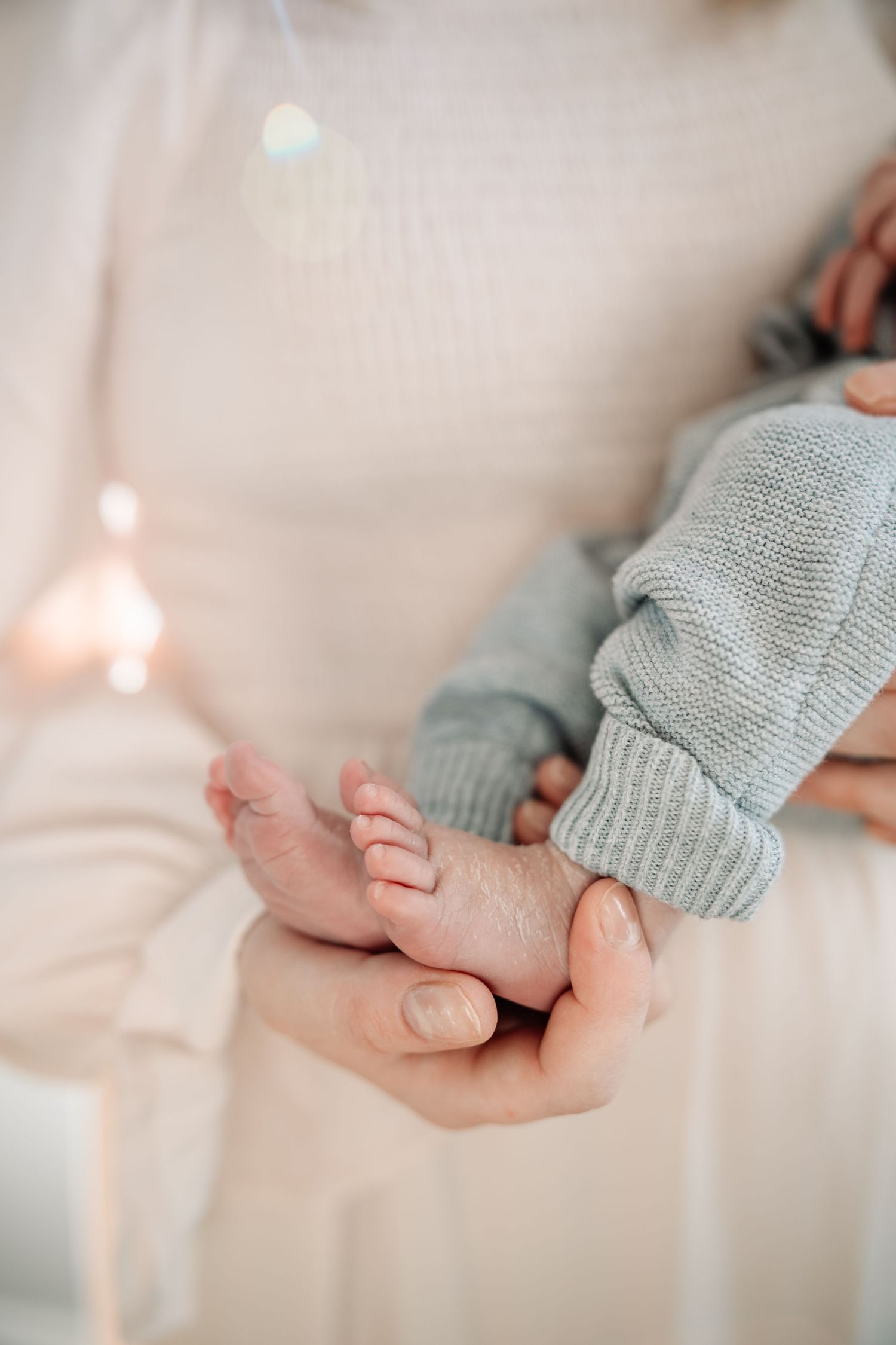 A mother gently holds her newborn baby's tiny feet in her hands, dressed in a soft gray knit outfit.