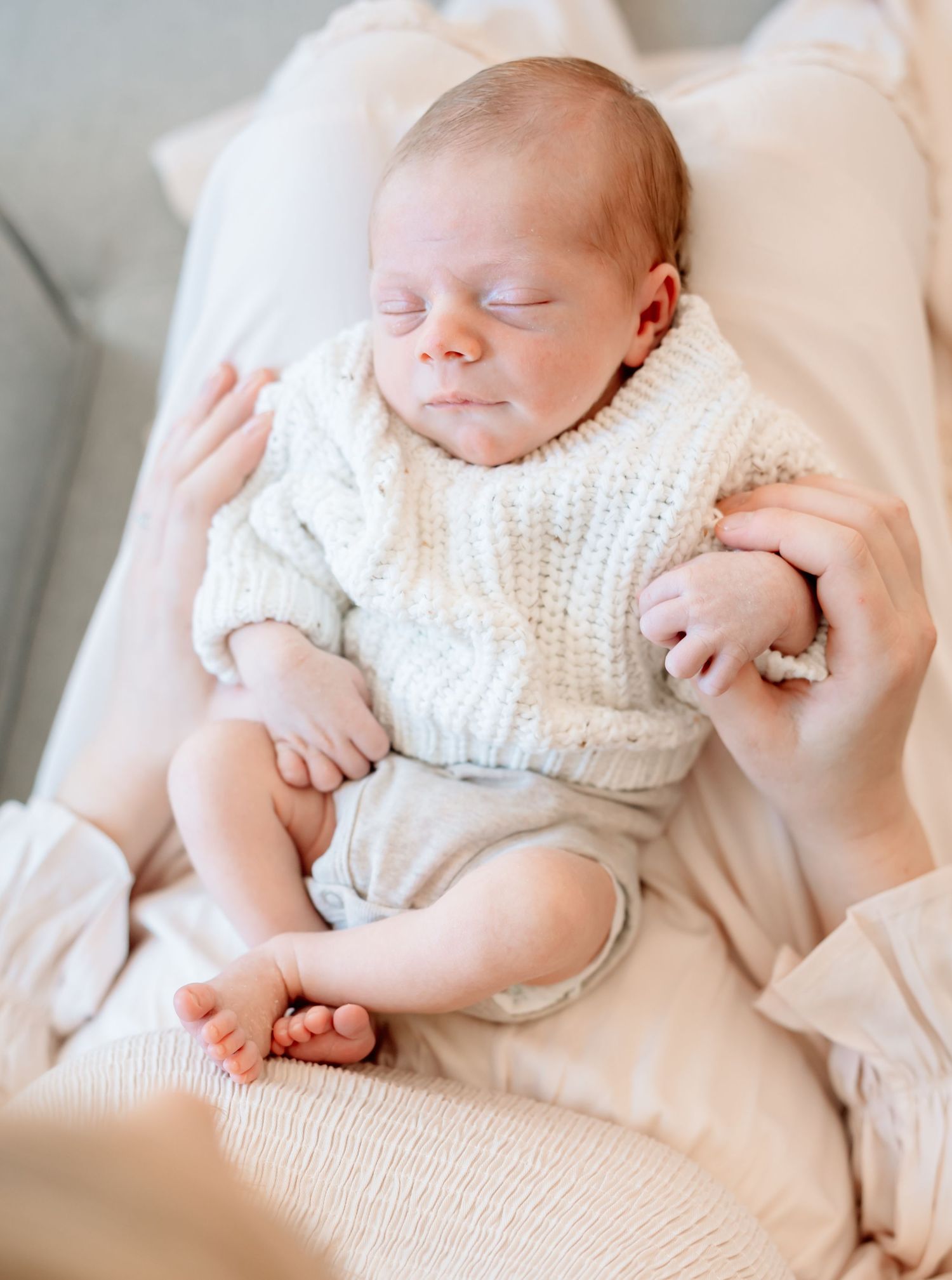 Peaceful newborn baby sleeping in white cable-knit sweater, cradled in parent's arms on soft pink blanket.