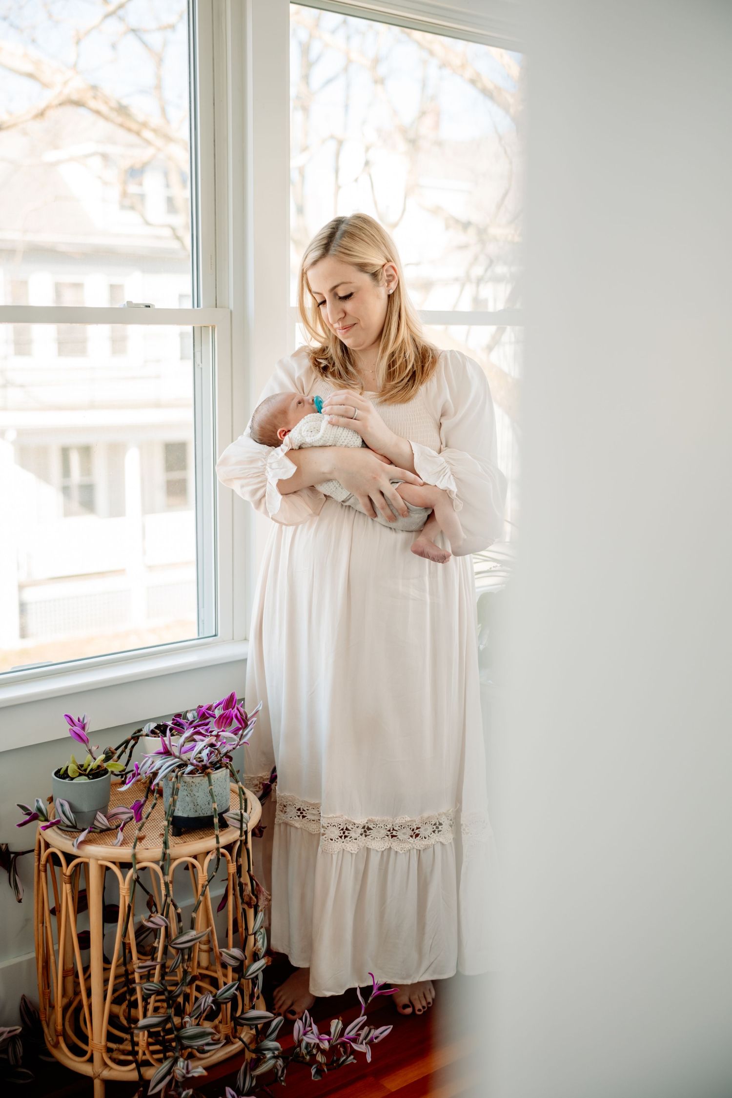 Blonde woman in white dress holding newborn baby by a sunlit window with plants nearby.