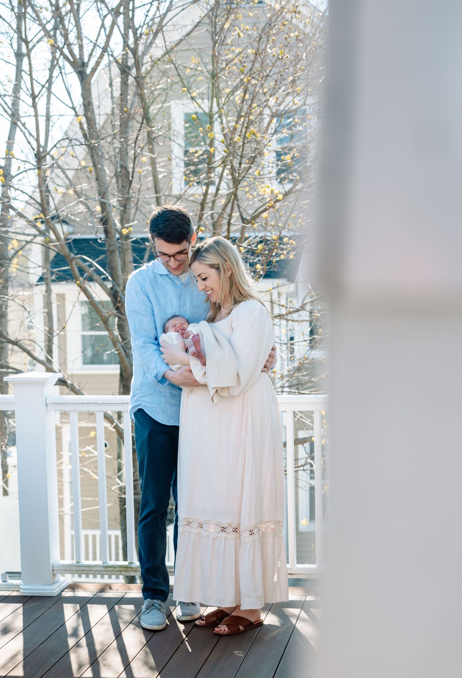 A couple stands on a porch gazing lovingly at their newborn baby, dressed in light blue and white.