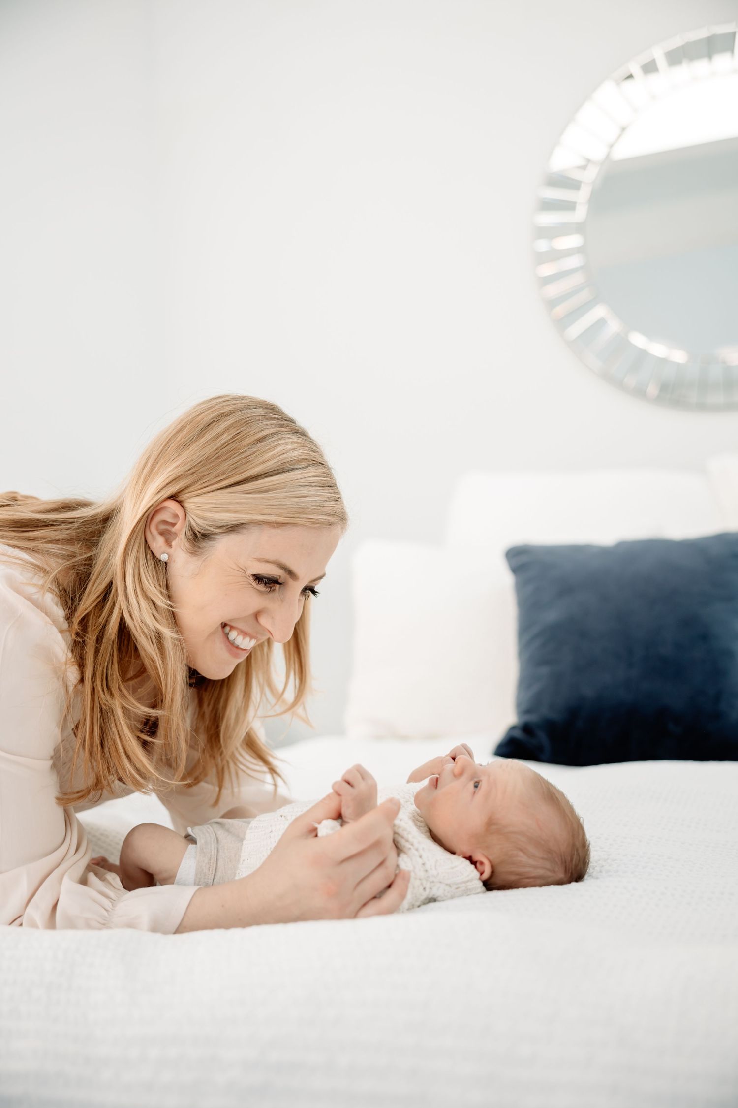 Smiling blonde mother lying on white bed interacting with her newborn baby in a bright, modern bedroom.