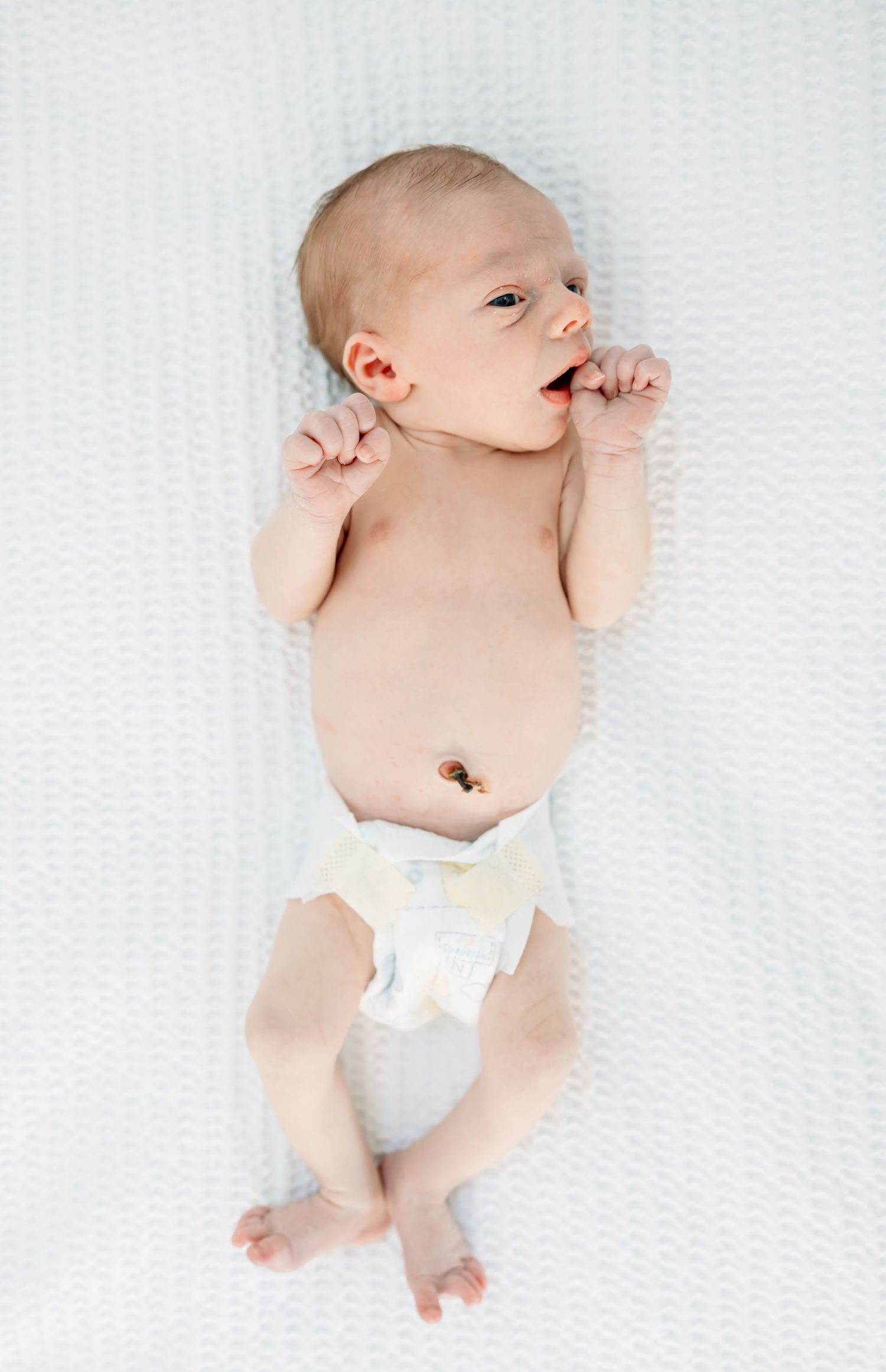 Newborn baby lying on white blanket wearing diaper with umbilical cord stump visible.