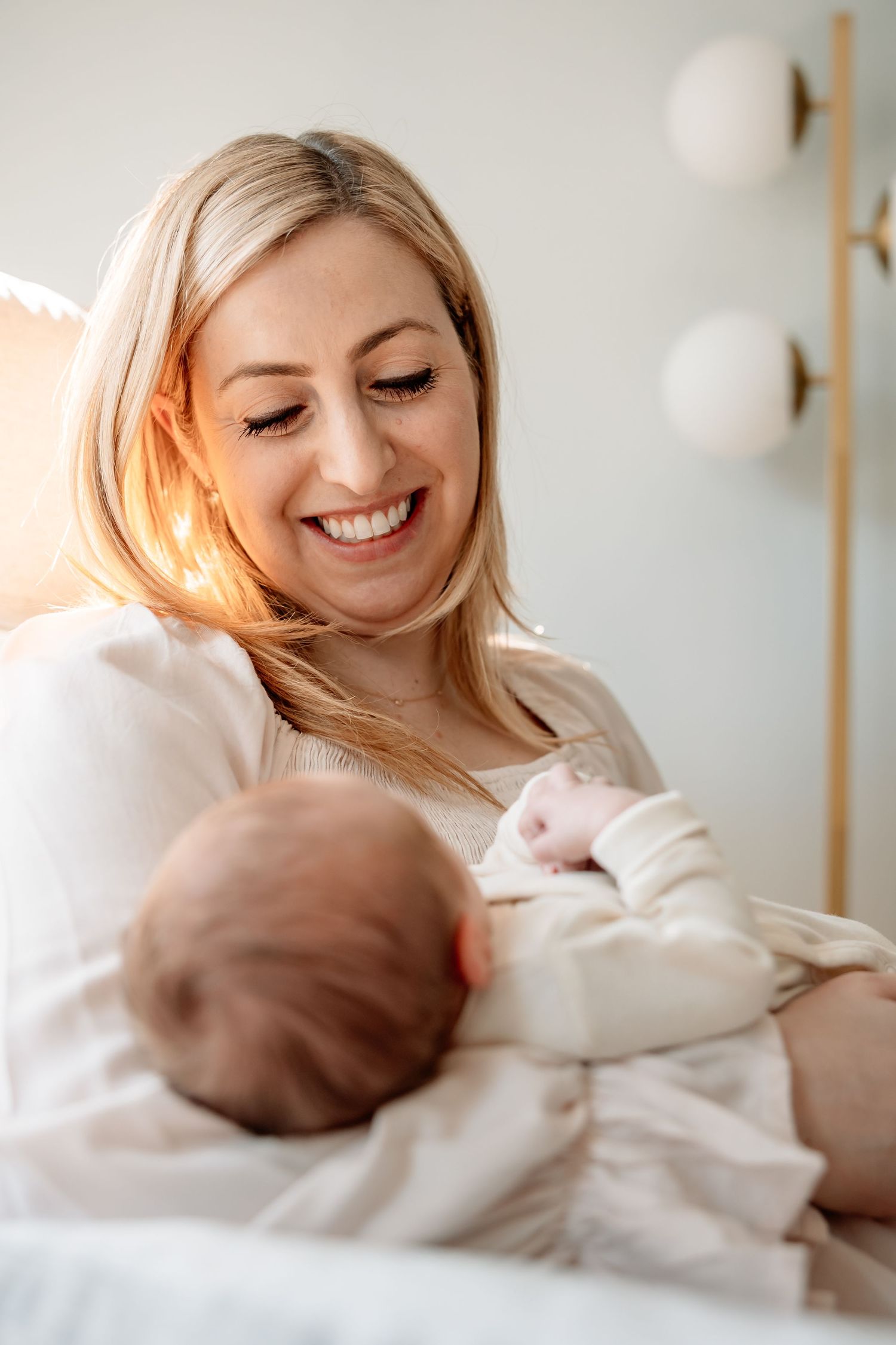 Smiling blonde mother holding and looking at her newborn baby in bright room.
