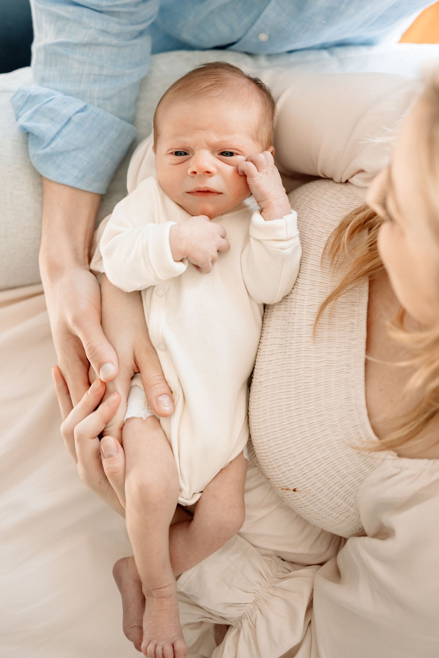 Newborn baby in white onesie lying on bed, cradled by two parents hands, looking up with curious expression.