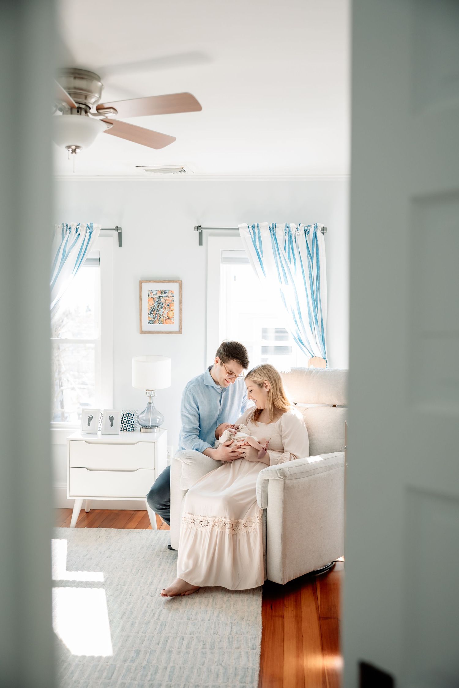 A couple sits together in a bright nursery, lovingly gazing at their newborn baby in a cozy white chair.