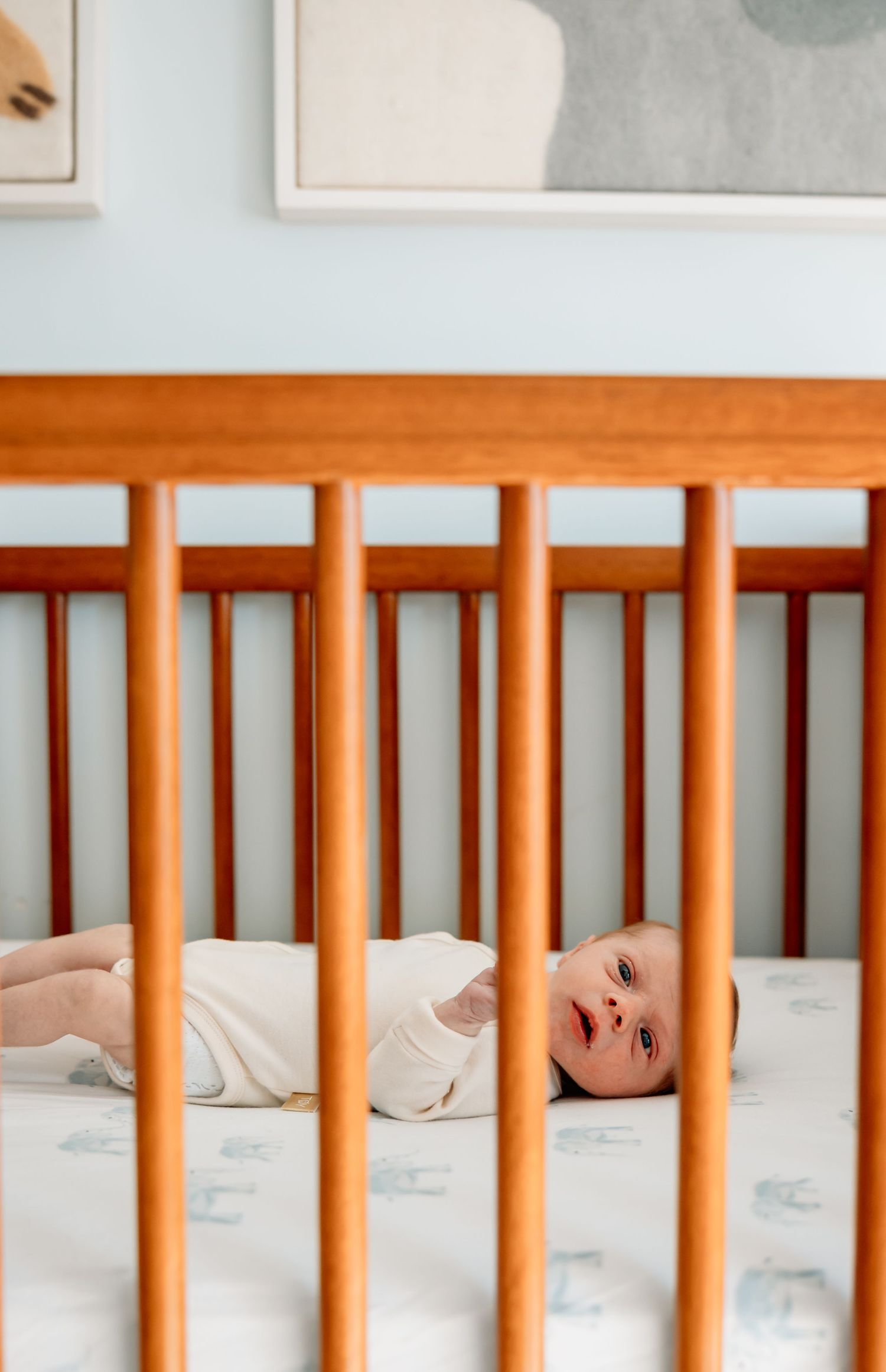 A peaceful newborn baby lies awake in a wooden crib with soft blue nursery decor in the background.