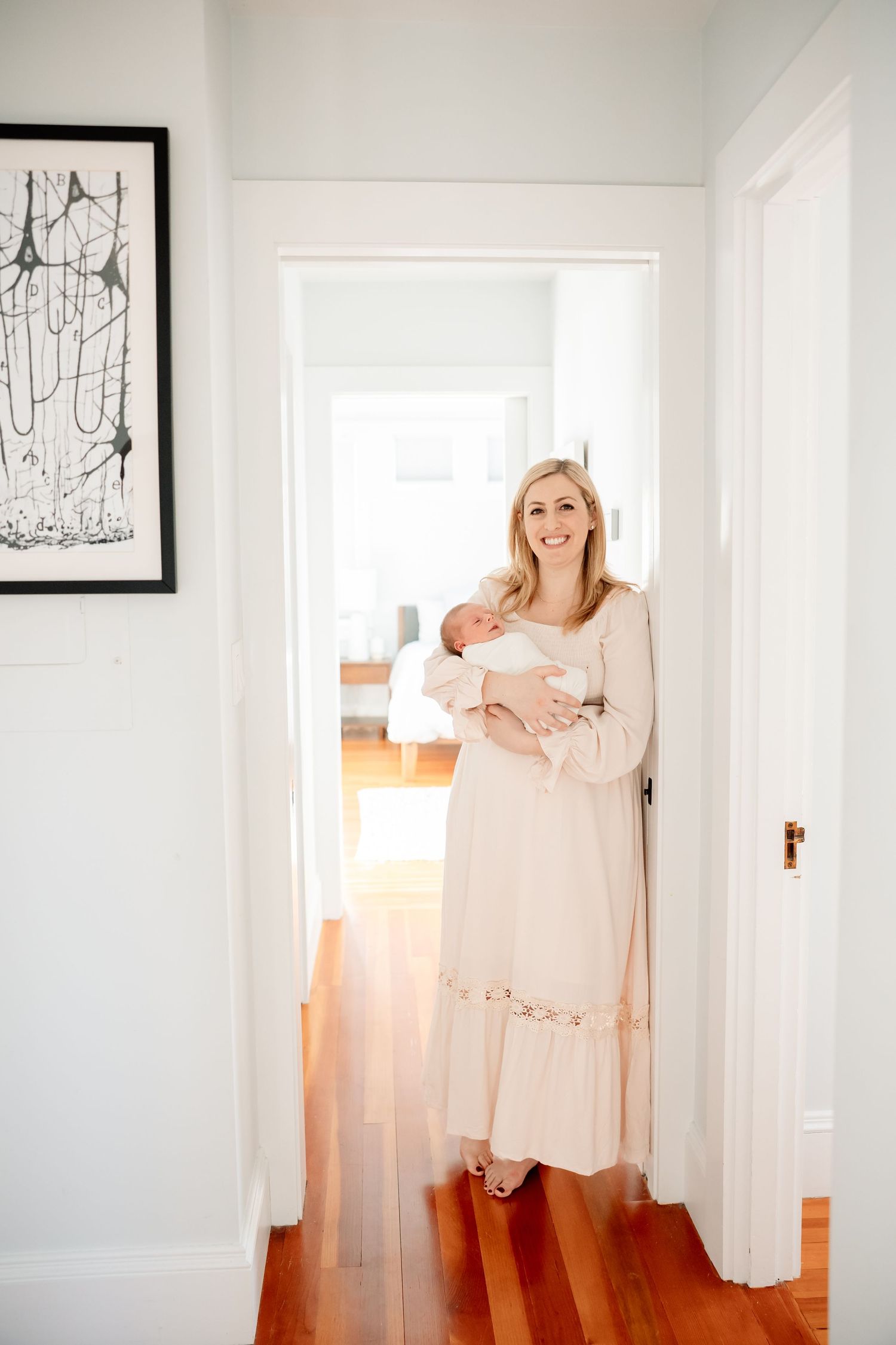 Smiling blonde mother in cream dress holds newborn baby in bright white hallway with hardwood floors.