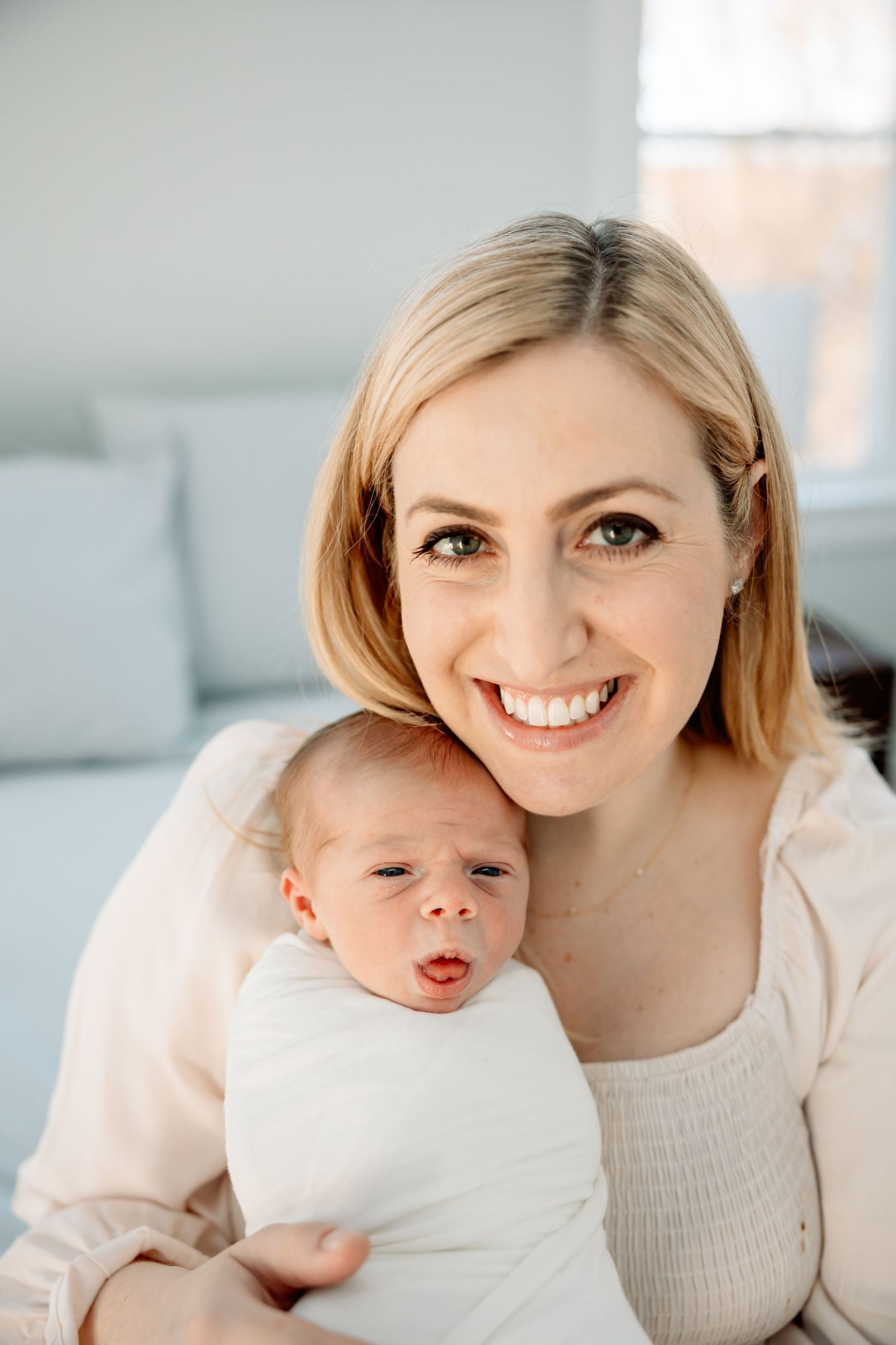 A smiling blonde mother holds her swaddled newborn baby close to her chest in a bright, airy room.