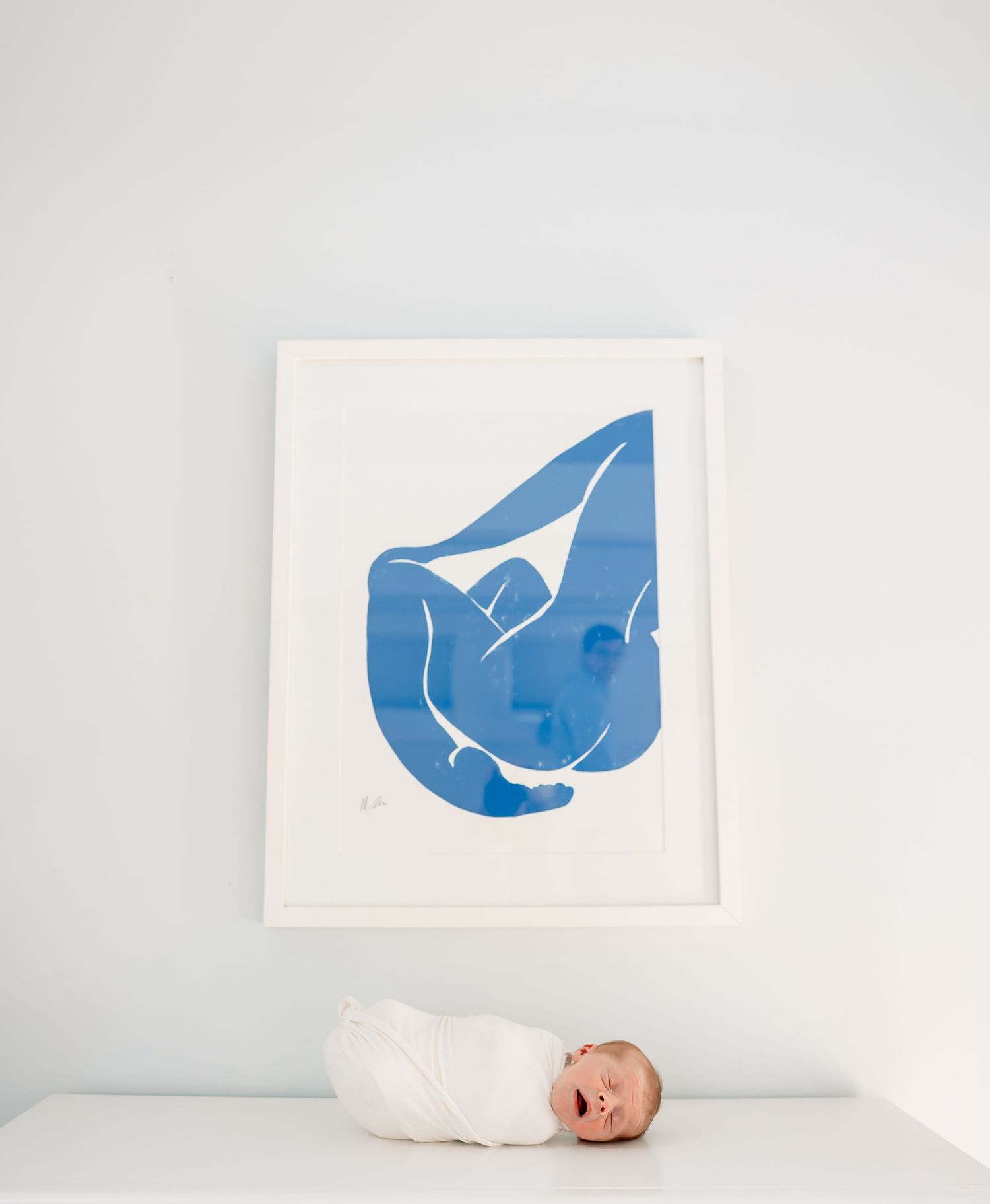 Swaddled newborn baby lies beneath framed blue abstract art print on white dresser in nursery.