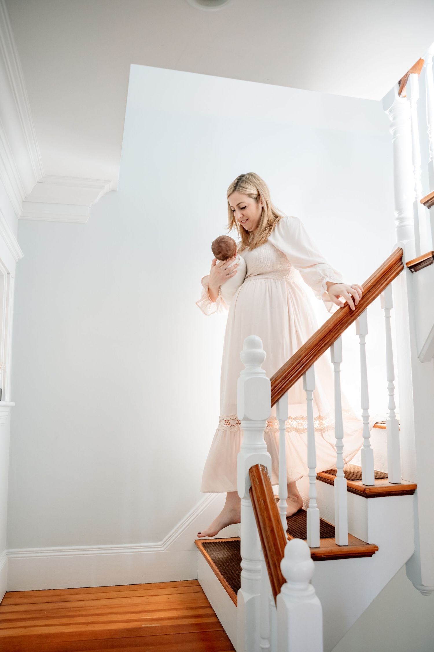 Blonde mother in flowing cream dress walks downstairs holding newborn baby on wooden staircase.
