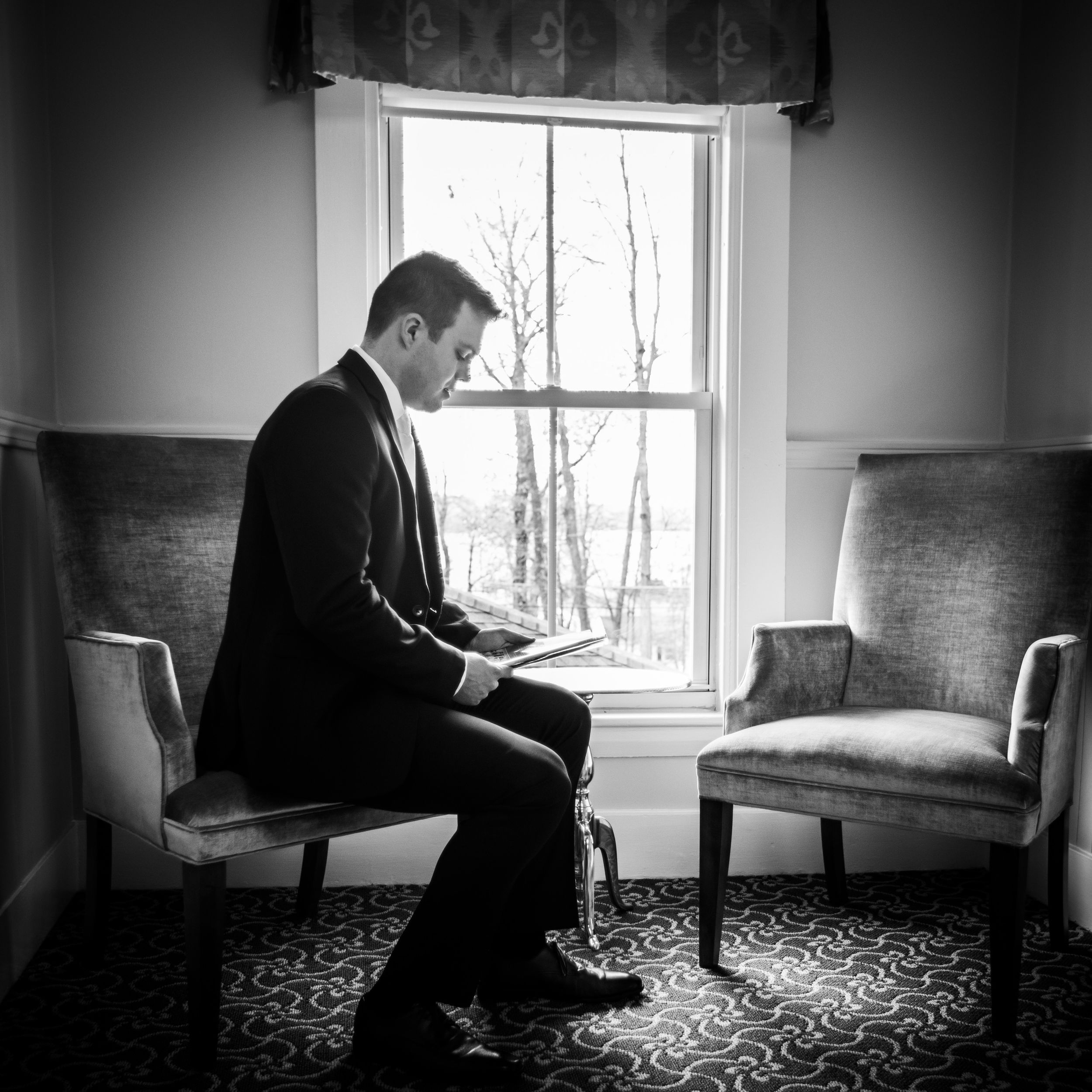 Black and white photo of a groom in a suit sitting by a window, reading notes before his wedding ceremony.
