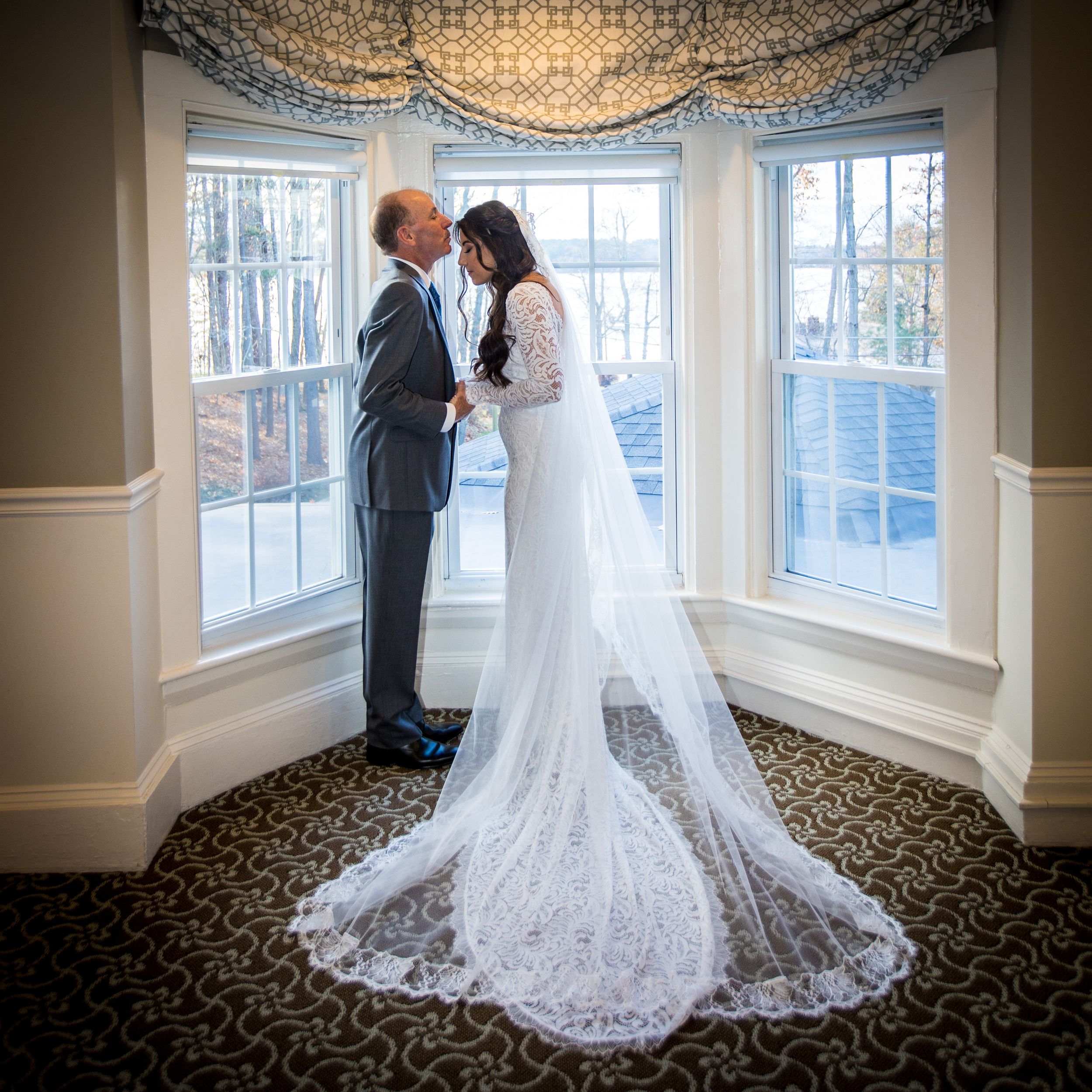 Bride and older man share tender moment by bay window, her lace cathedral veil trailing elegantly on patterned carpet.
