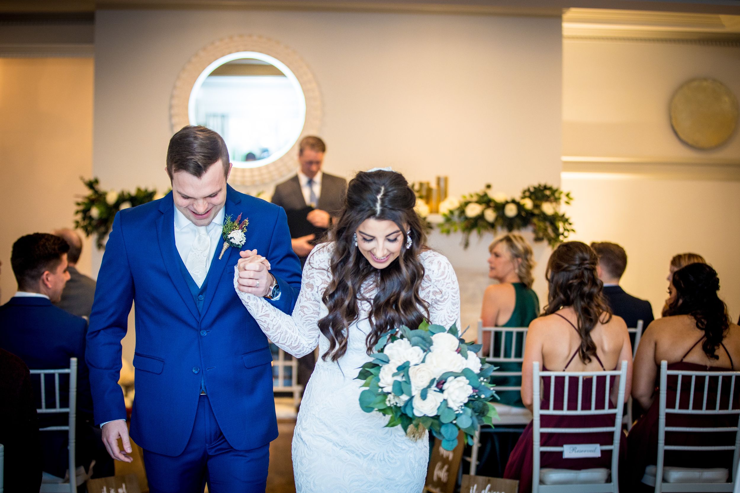 Newlyweds walk down aisle hand in hand, bride holding white bouquet, guests seated in decorated wedding venue.